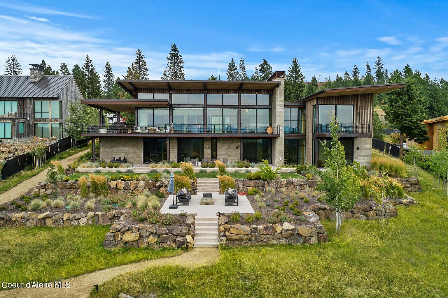 This is a rear view of a modern, multi-level home with extensive landscaping. The house features large windows, balconies, and a stone patio with outdoor seating and a fire pit. The landscaping includes terraced rock walls, ornamental grasses, and a winding path leading to the house, creating a luxurious and inviting outdoor space.