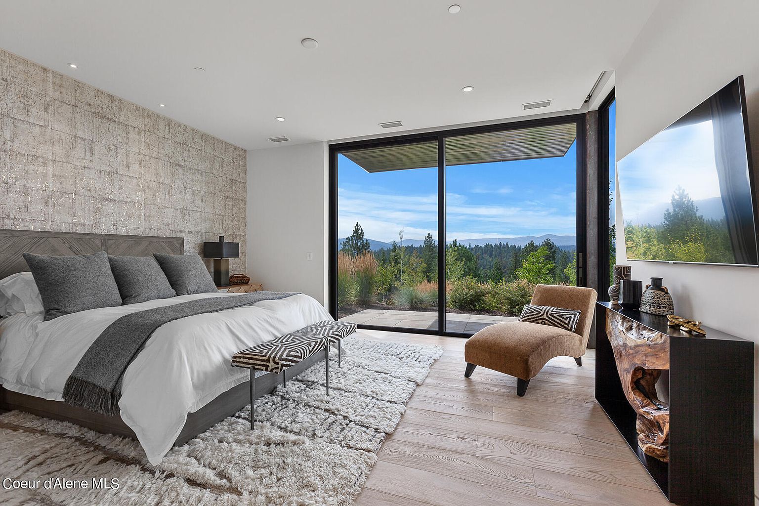 This primary bedroom features a modern design with a textured accent wall behind the bed. Large windows offer stunning views of the surrounding landscape, bringing natural light into the space. The room is furnished with a bed, bench, chaise lounge, and a sleek console table with a television, creating a luxurious and comfortable atmosphere.