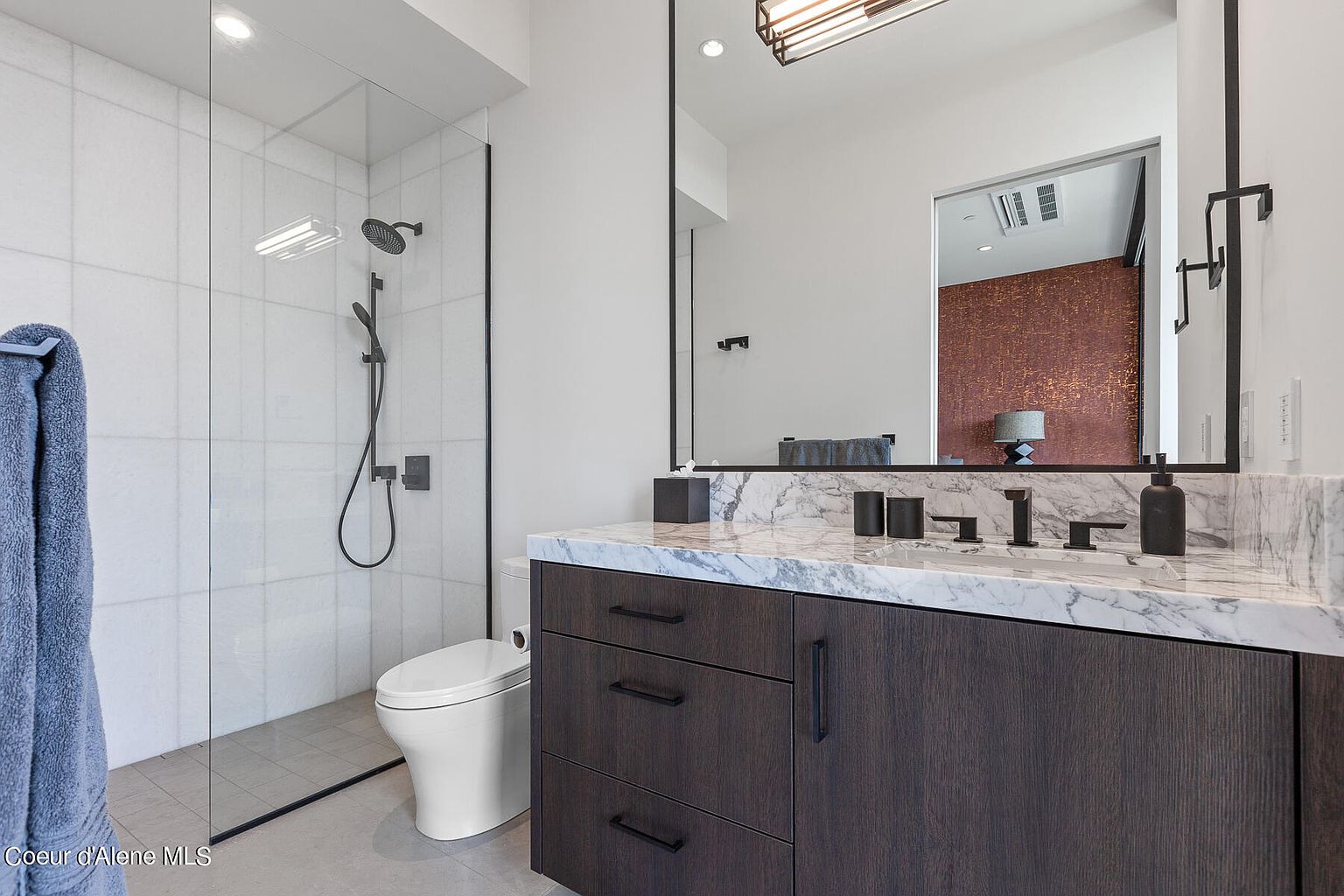 This is a primary bathroom featuring a modern design with a walk-in shower enclosed in glass, a toilet, and a vanity with a marble countertop. The vanity has dark wood cabinetry with sleek, black hardware. A large mirror hangs above the vanity, reflecting a glimpse into another room with textured walls, and the overall aesthetic is clean and luxurious.