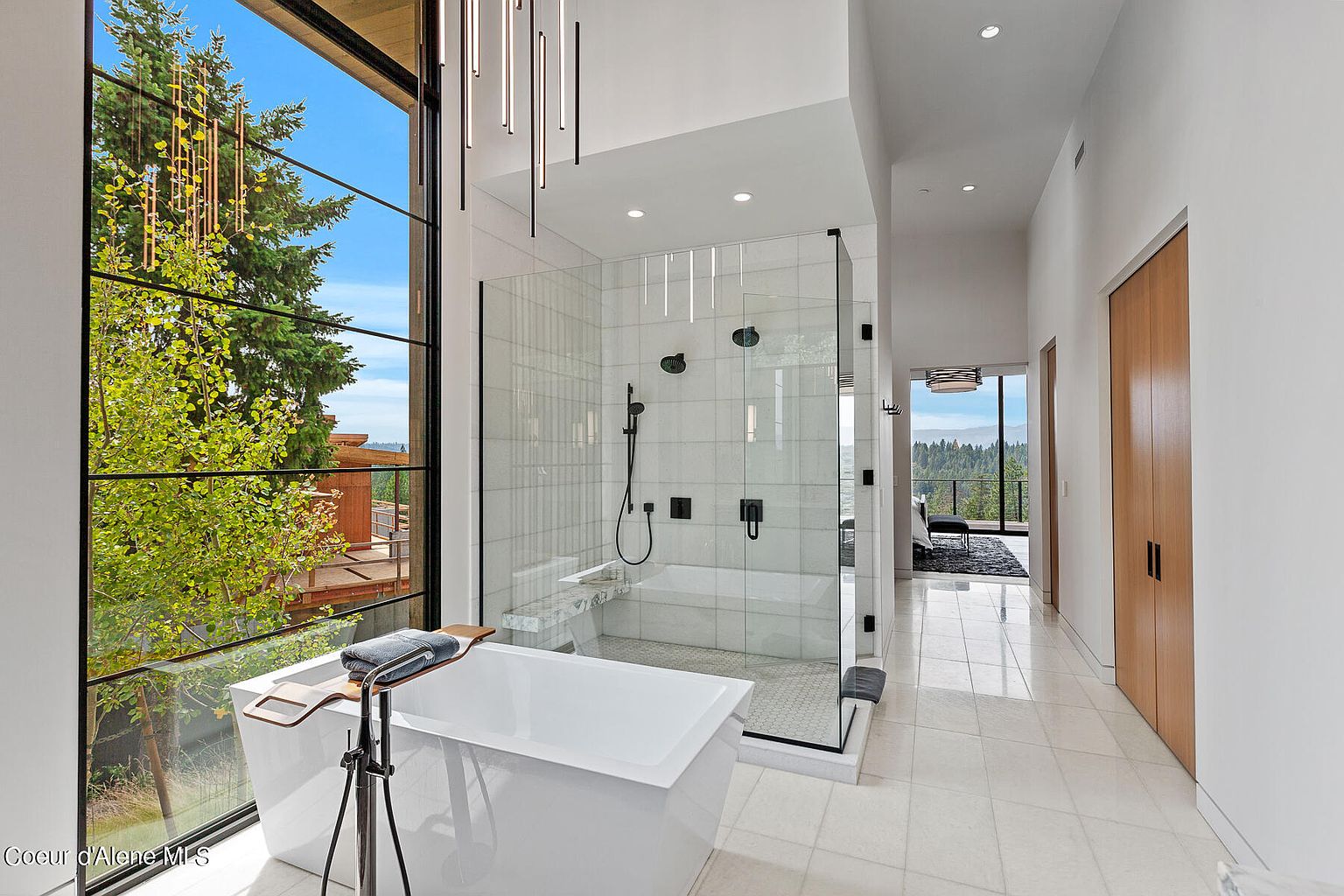 This is a luxurious primary bathroom featuring a freestanding white bathtub positioned next to a large window with a view of the outdoors. A glass-enclosed shower with white tile and black fixtures is also present. The bathroom has a modern design with white tile flooring and a hallway leading to another room.