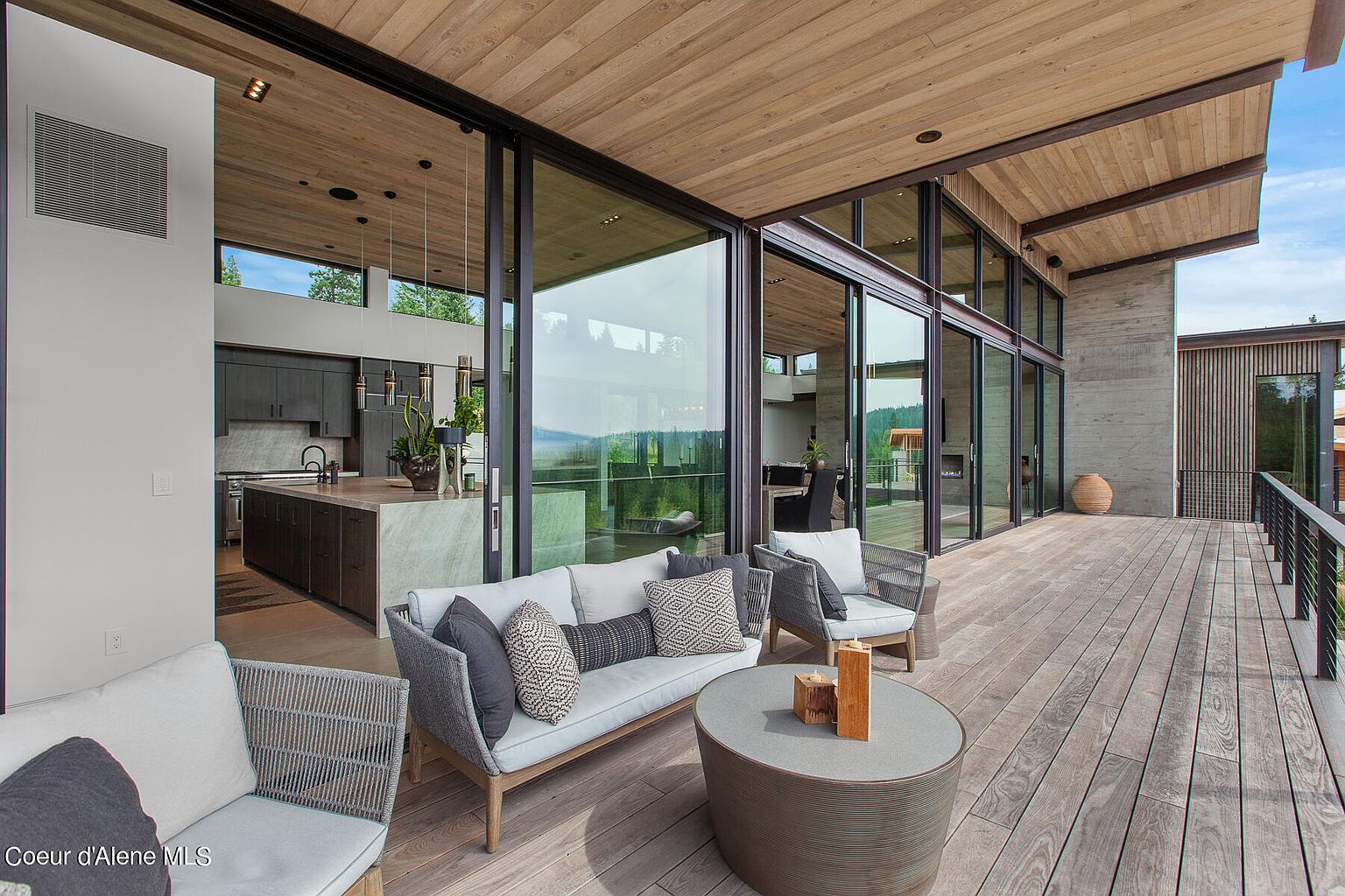 This image showcases a luxurious outdoor patio or deck area, seamlessly blending indoor and outdoor living. The space features comfortable seating arrangements, including a sofa and chairs, arranged around a stylish coffee table. Large sliding glass doors provide access to the interior, offering stunning views of the surrounding landscape and creating an inviting atmosphere for relaxation and entertainment.