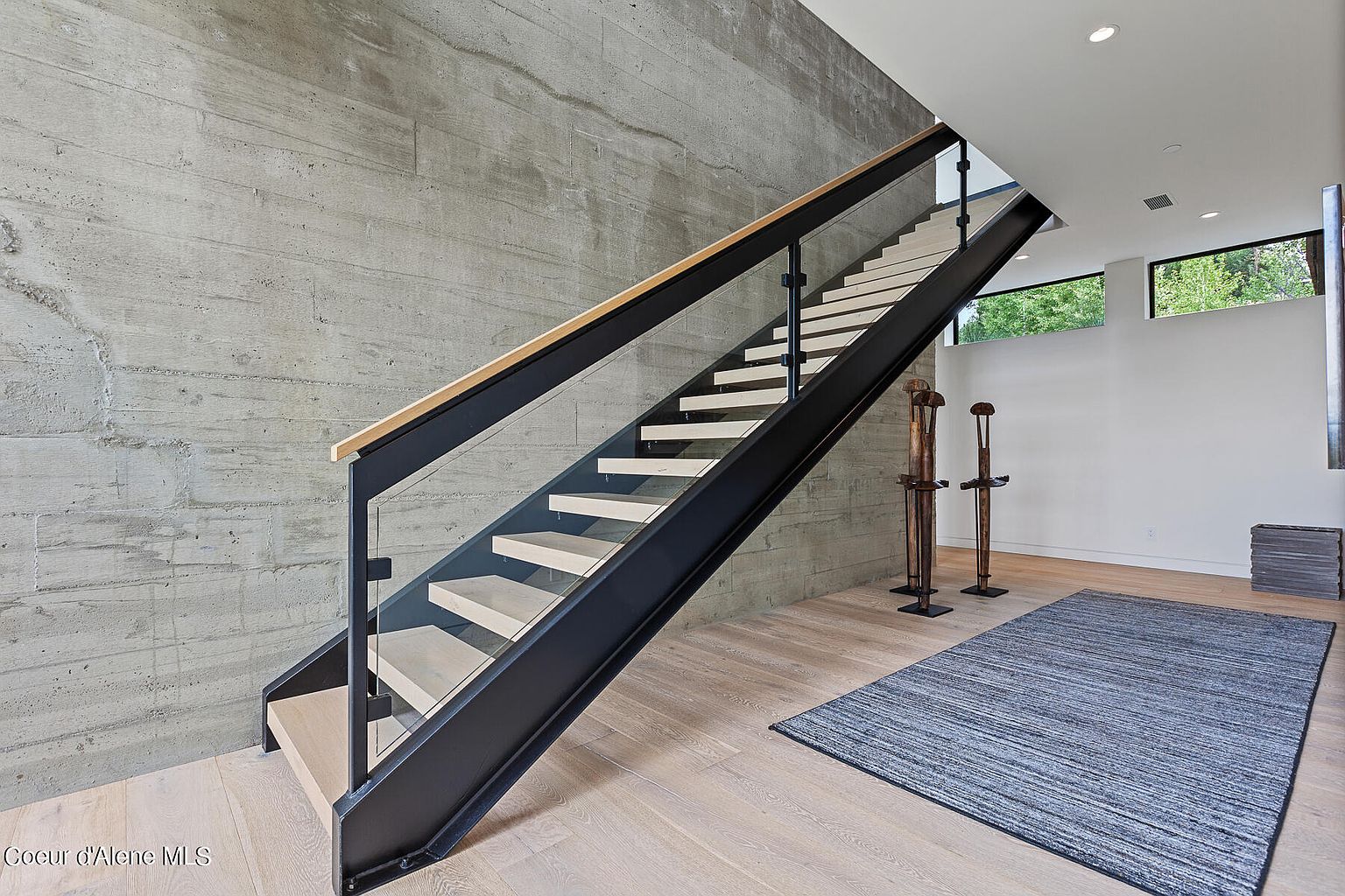 This interior shot showcases a modern staircase with light wood treads and a black metal frame, complemented by a glass railing and a light wood handrail. The staircase ascends against a textured concrete wall, while the adjacent space features minimalist decor, including two wooden sculptures and a blue rug on light wood flooring. The overall impression is one of contemporary design and clean lines.
