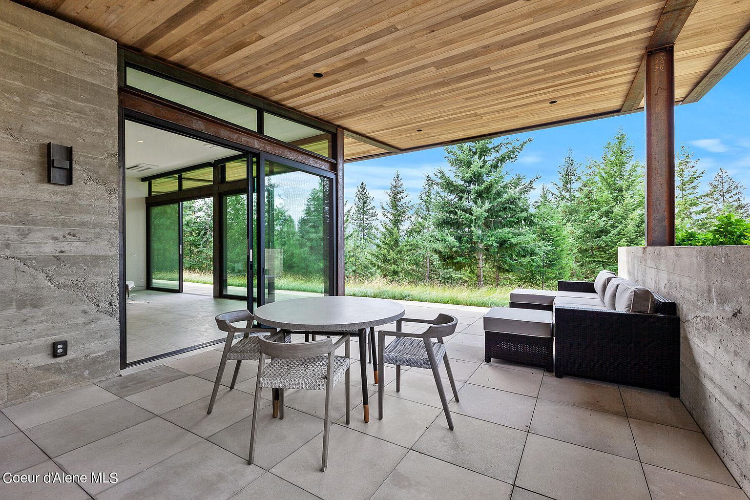 This image showcases a modern patio area with a round table and chairs, along with a comfortable outdoor sofa. The patio features a sleek, tiled floor and a wooden ceiling, seamlessly blending indoor and outdoor living. Large sliding glass doors open to a view of lush greenery and trees, creating a serene and inviting atmosphere.