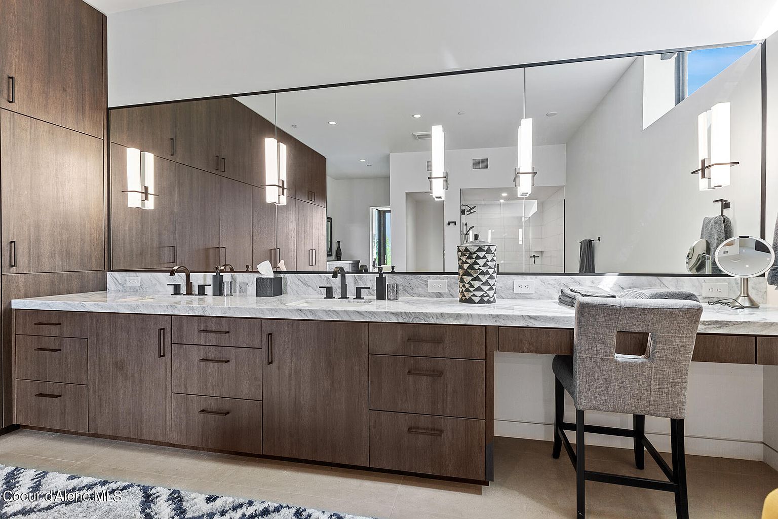 This is a primary bathroom featuring a double vanity with a marble countertop and dark wood cabinetry. A large mirror spans the length of the vanity, reflecting pendant lighting and a glimpse into the rest of the bathroom. A makeup vanity with a chair is on the right side of the image.