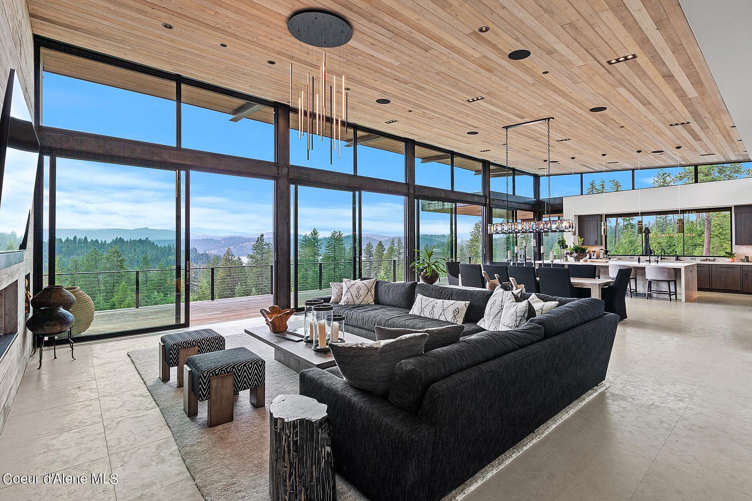 This expansive living room boasts floor-to-ceiling windows offering panoramic views of the surrounding landscape. The open-concept design seamlessly integrates the living area with the dining and kitchen spaces, creating a cohesive and inviting atmosphere. A large sectional sofa anchors the room, complemented by modern lighting fixtures and a natural wood ceiling, enhancing the home's contemporary yet warm aesthetic.