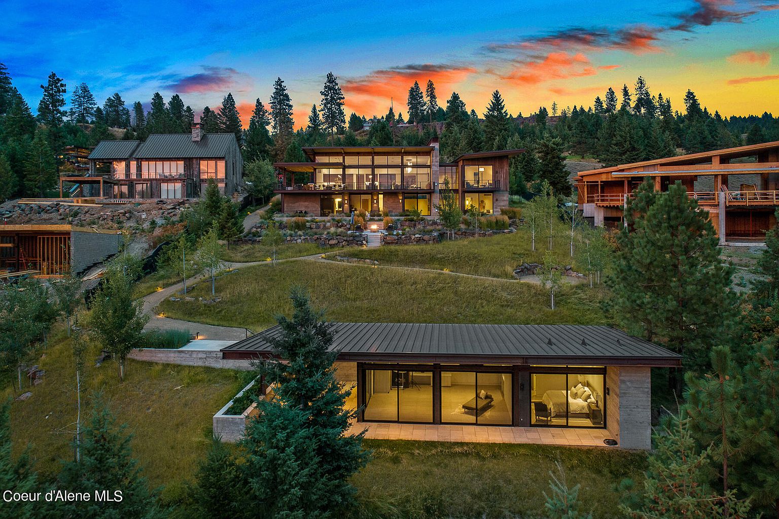 This aerial view showcases a collection of modern homes nestled into a hillside landscape at sunset. The architecture features clean lines, large windows, and natural materials, blending seamlessly with the surrounding trees and greenery. The scene evokes a sense of luxury, privacy, and connection with nature.