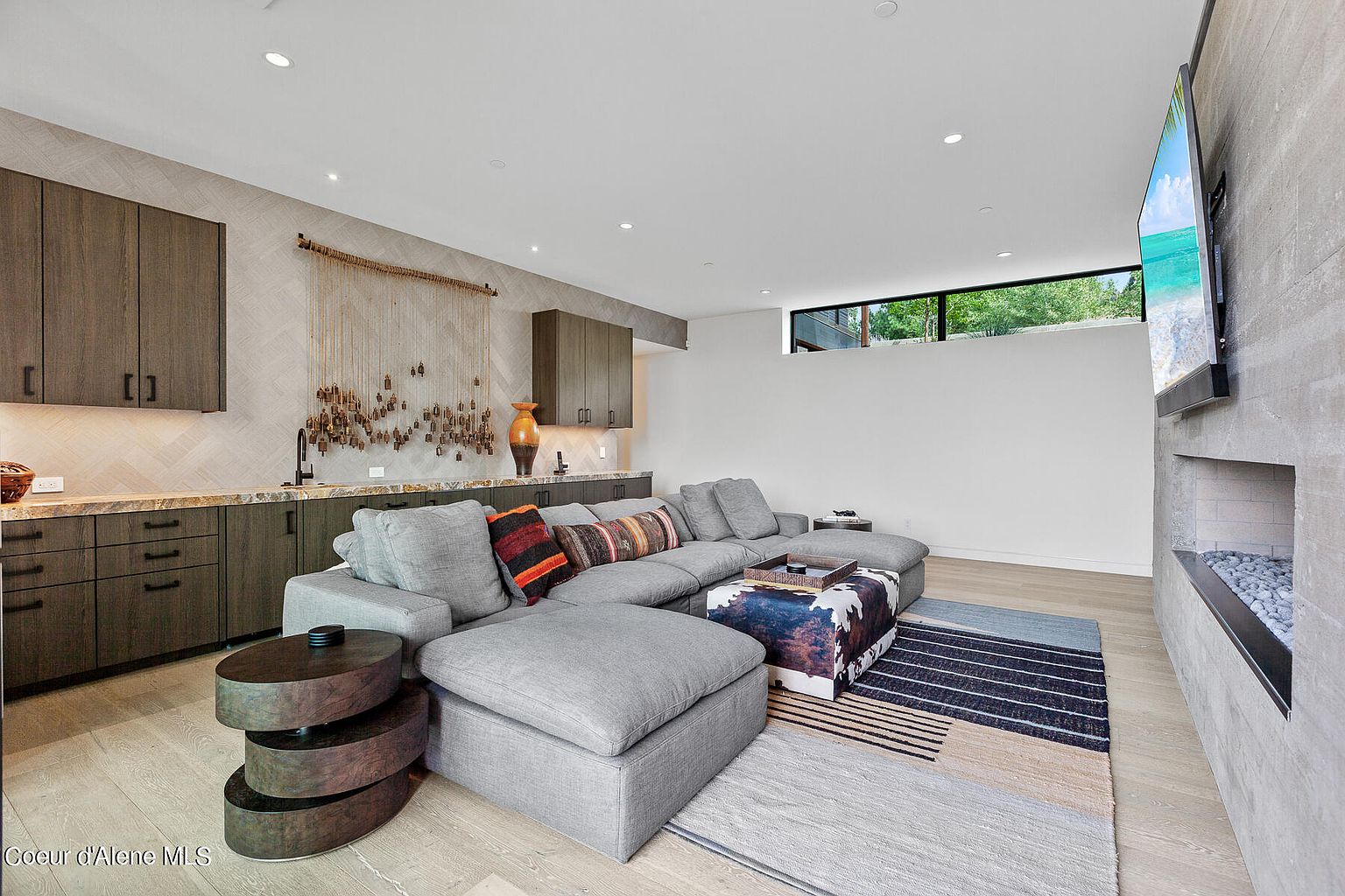 This is an interior shot of a modern living room featuring a large, sectional sofa in a neutral gray tone, complemented by decorative pillows and a unique ottoman. The room includes custom cabinetry with a wet bar, a contemporary fireplace with a television mounted above, and stylish area rugs layered on light wood flooring. The perspective is from a low angle, showcasing the depth and design elements of the space.