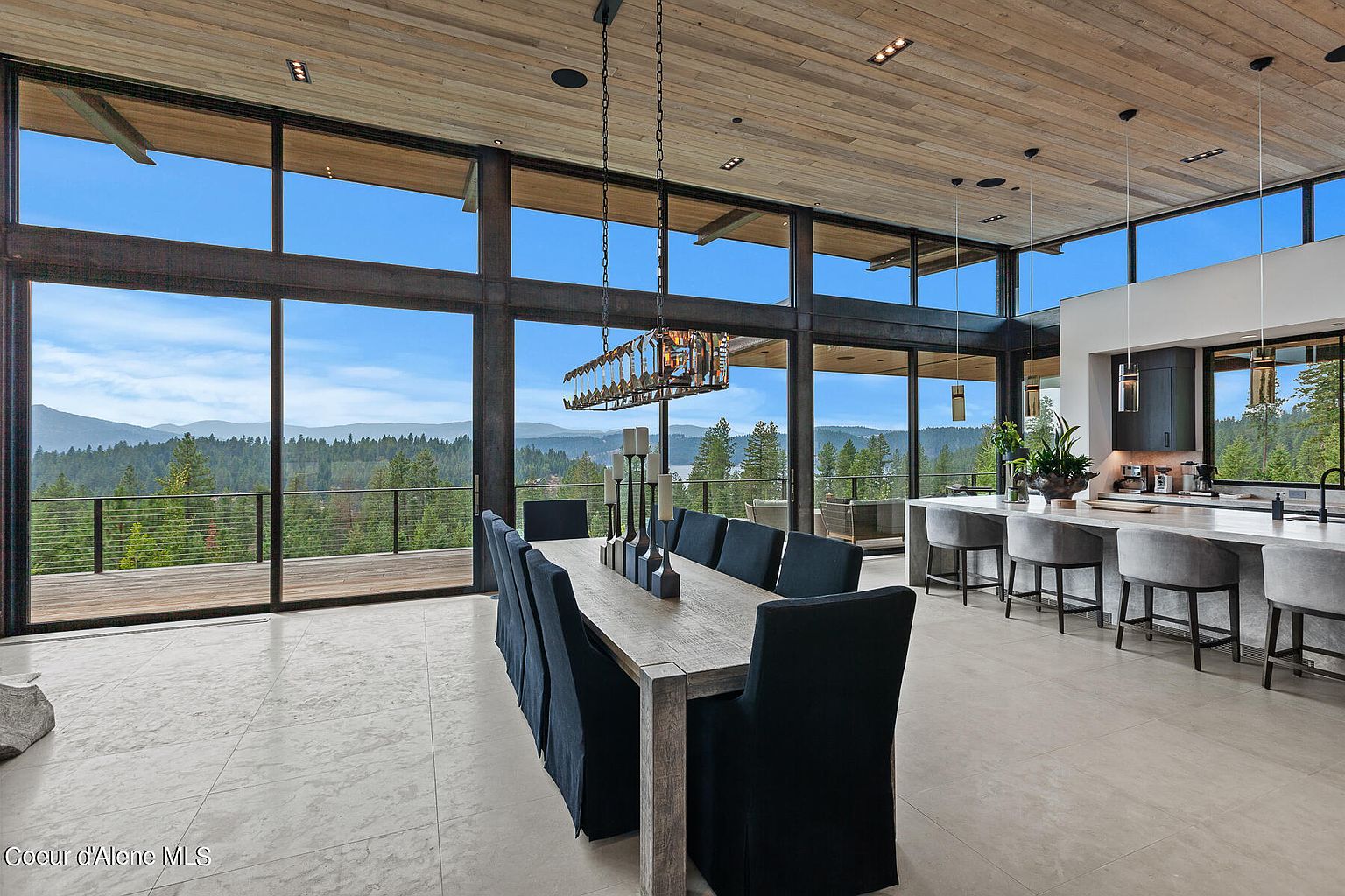 This is a spacious dining room featuring a long wooden table with blue upholstered chairs, set beneath a modern chandelier. Large windows offer panoramic views of the surrounding landscape, bringing natural light into the room. The open-concept design seamlessly connects the dining area to the kitchen, creating an inviting and luxurious atmosphere.