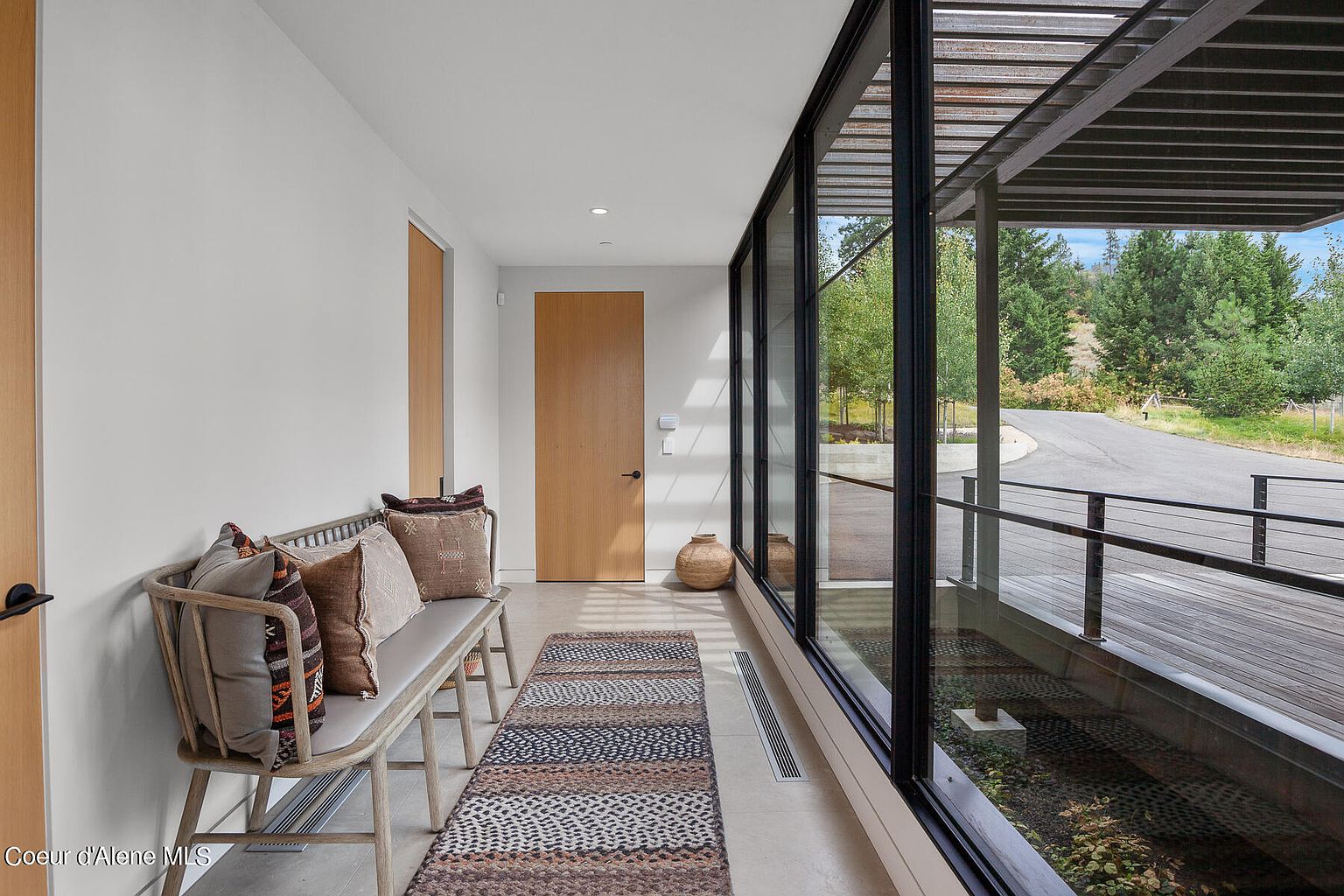 This interior shot showcases a modern hallway with a bench adorned with decorative pillows, a patterned rug, and sleek wooden doors. A wall of windows provides ample natural light and a view of the exterior landscape, creating a seamless indoor-outdoor transition. The design emphasizes clean lines and a minimalist aesthetic.