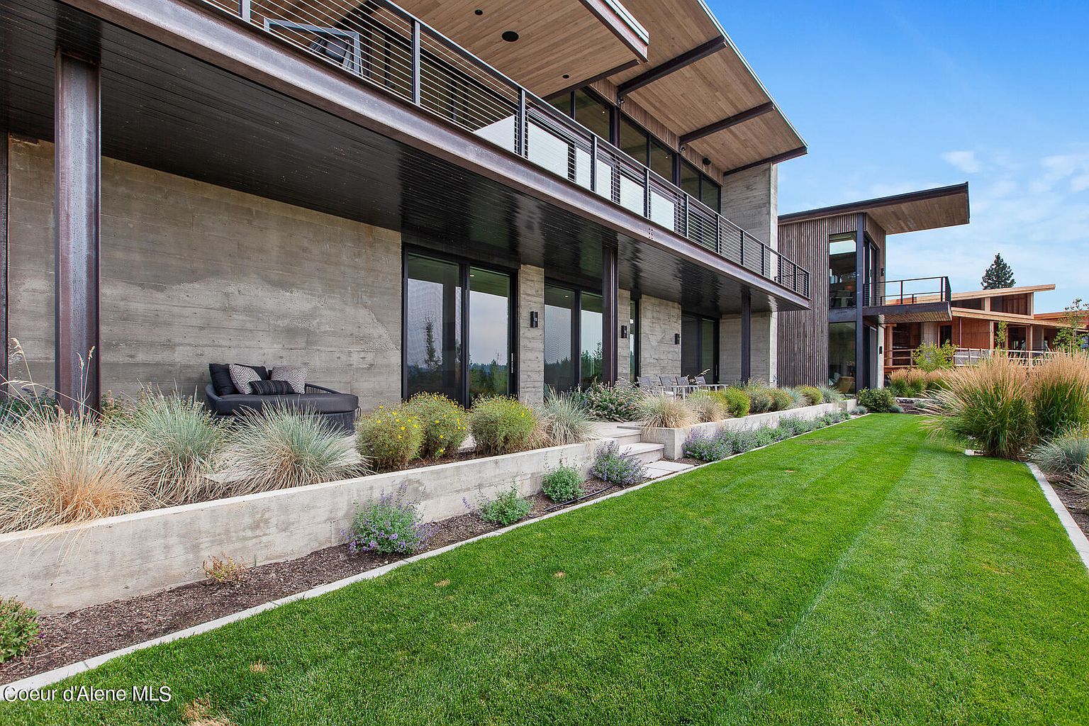 This image showcases the exterior of a modern home, focusing on the meticulously maintained yard and garden area. The architecture features a concrete base with dark-framed windows and a balcony above. The lush green lawn is neatly striped, complemented by a well-designed garden bed with various plants and shrubs, creating an inviting outdoor space.