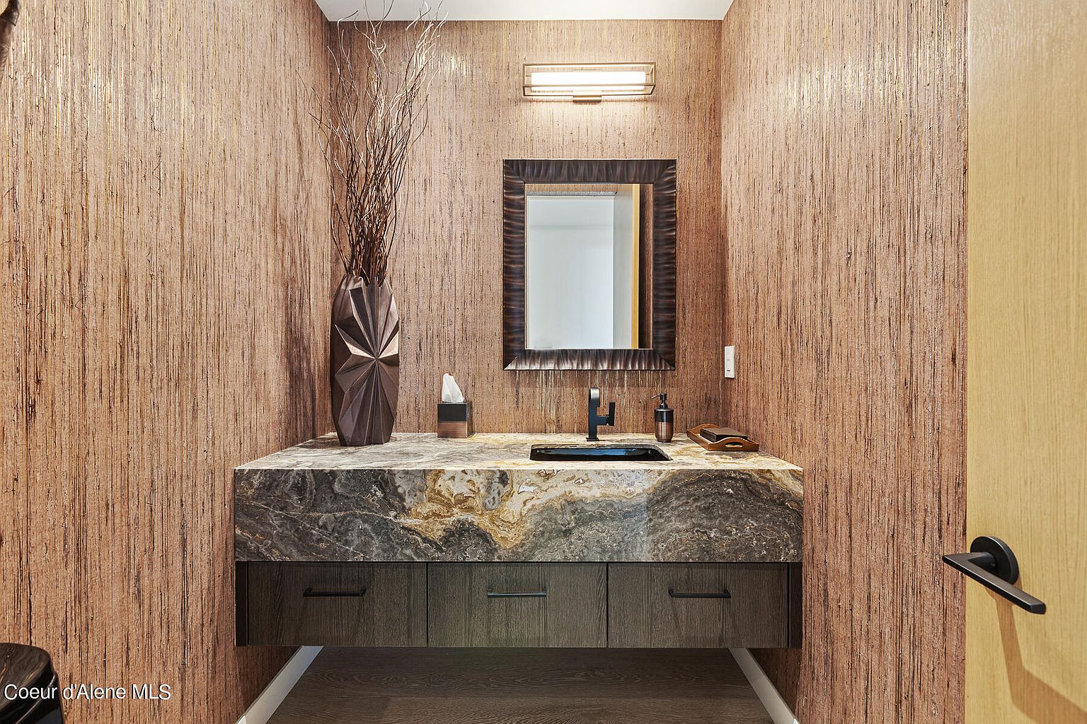 This is a stylish guest bathroom featuring a unique marble countertop with a dark vessel sink and matte black faucet. The walls are covered in textured wallpaper, complemented by a framed mirror and modern light fixture. A decorative vase with branches adds a touch of elegance, and the dark wood cabinetry provides ample storage.