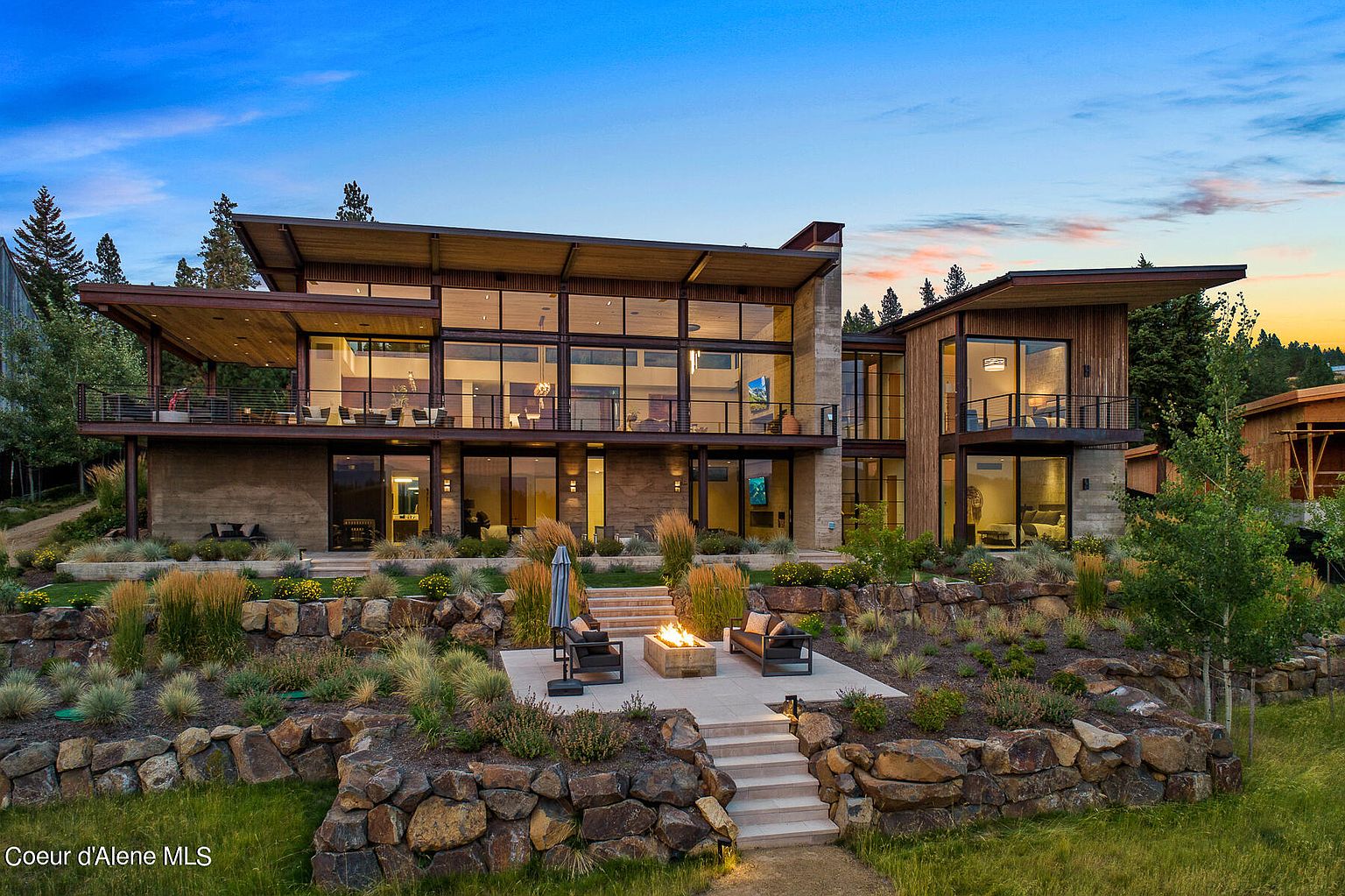This image showcases the rear exterior of a modern, multi-level home with expansive windows and a tiered landscape. A patio area with a fire pit and seating is nestled within the stone retaining walls and lush greenery, creating an inviting outdoor living space. The architectural design emphasizes clean lines and a seamless integration with the natural surroundings.