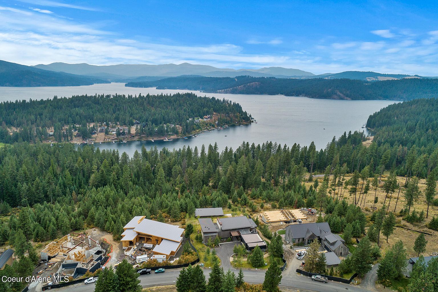 This aerial shot showcases a collection of modern homes nestled amidst a lush forest, with a serene lake and distant mountains providing a stunning backdrop. The properties feature contemporary architectural designs, some still under construction, highlighting the exclusive and developing nature of the real estate. The scene conveys a sense of luxury, privacy, and natural beauty, ideal for high-end buyers seeking a secluded retreat.