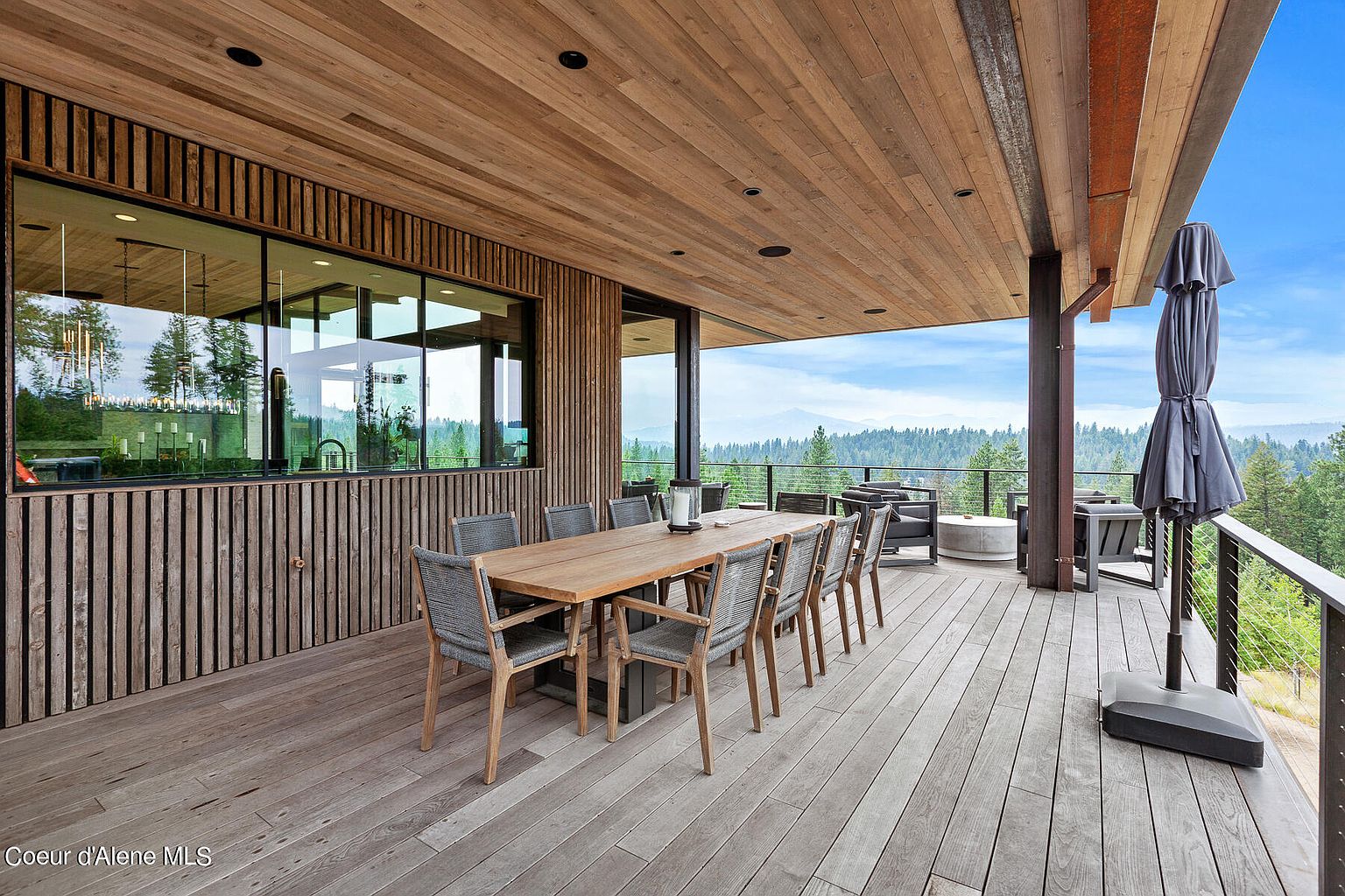 This image showcases a spacious outdoor deck area, featuring a large wooden dining table with seating for ten. The deck is constructed with light-colored wood planks and offers stunning views of the surrounding mountains and forest. An umbrella provides shade, and modern outdoor furniture adds to the luxurious feel of the space.