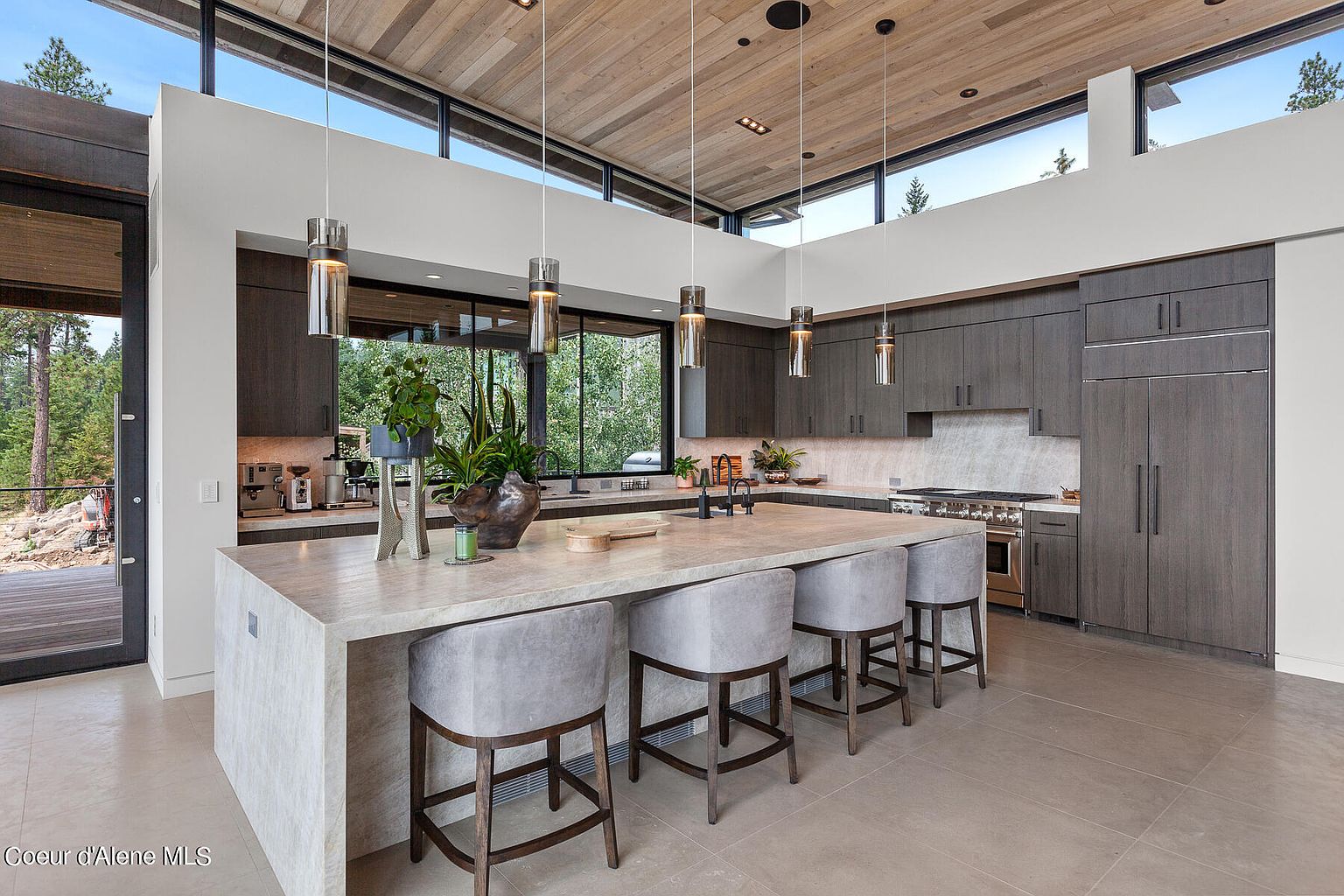 This is a modern kitchen with dark wood cabinetry, stainless steel appliances, and a large island with a light-colored countertop and seating for four. The kitchen features large windows offering a view of the surrounding trees, and pendant lighting hangs above the island. The overall impression is one of contemporary elegance and functionality.