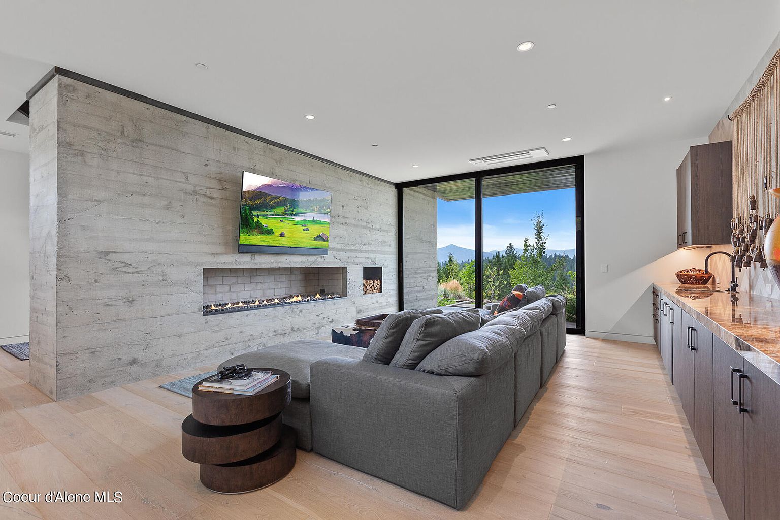 This is an interior shot of a modern living room featuring a large gray sectional sofa, a concrete accent wall with a linear fireplace and a mounted television. A round, tiered side table sits next to the sofa. Large windows offer a view of the outdoor landscape, and a wet bar area is visible on the right side of the frame. The room has a contemporary design with light wood flooring.