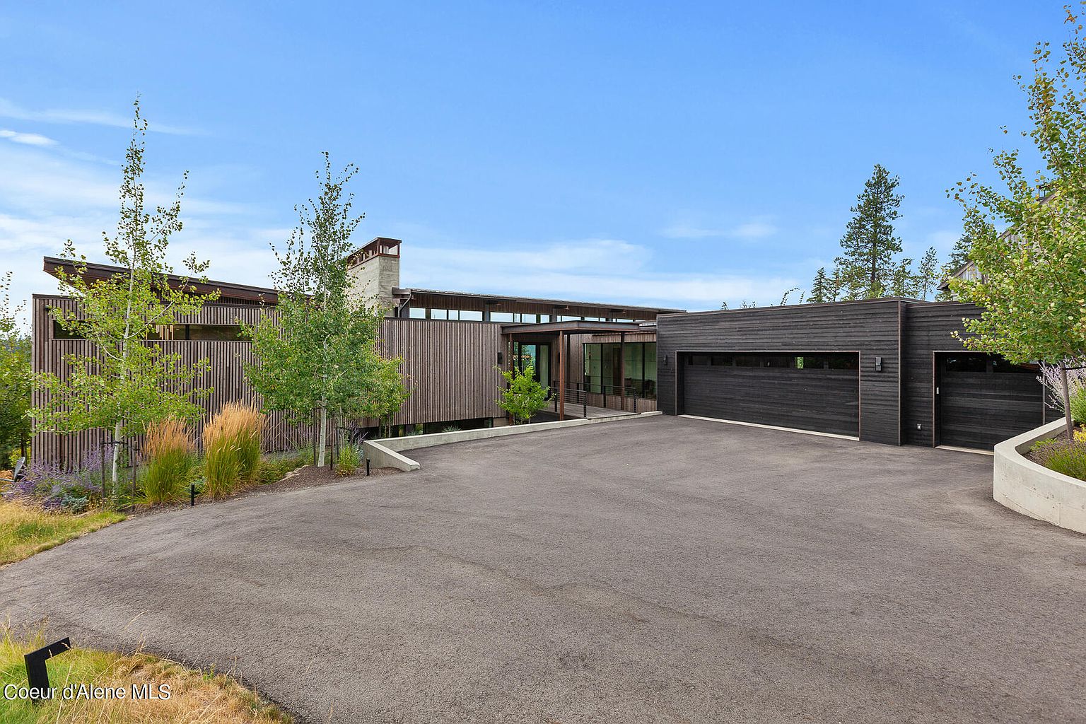 This is a front exterior view of a modern home featuring a combination of wood and dark siding. The house has a low-slung roofline and large windows, with a spacious driveway leading up to the attached garage. Landscaping includes trees and ornamental grasses, creating a natural and inviting entrance.