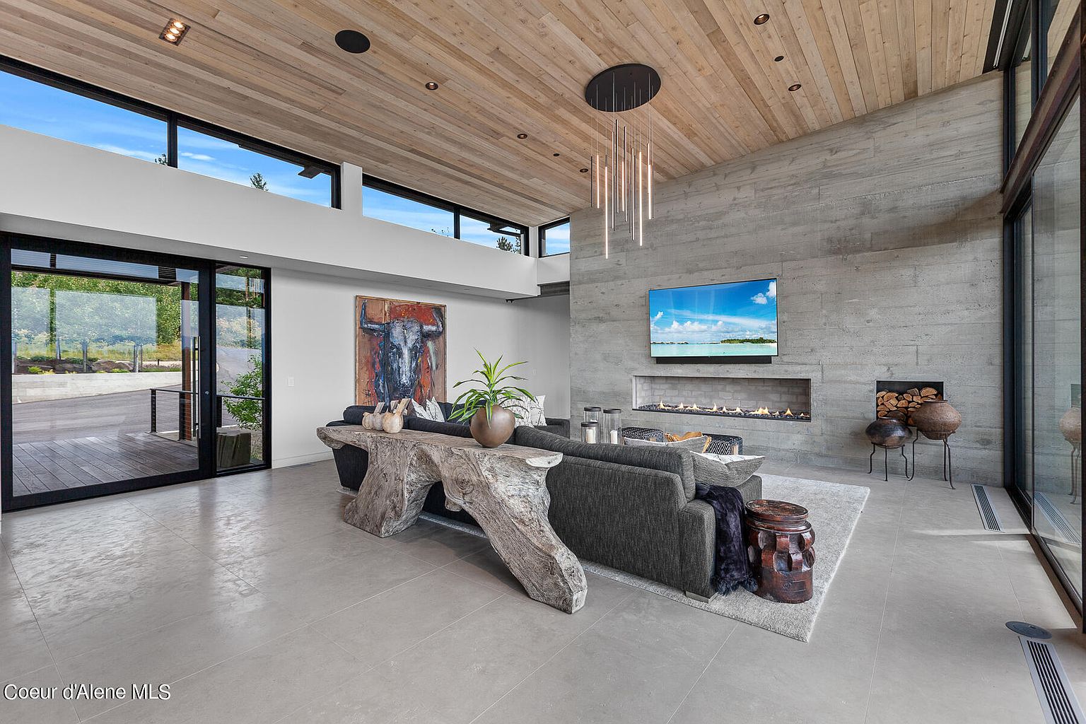 This is a modern living room featuring a concrete accent wall with a linear fireplace and a large television. The room has a high, wood-paneled ceiling with recessed lighting and a unique chandelier. A large gray sofa is complemented by a rustic wooden console table, and the room is filled with natural light from the expansive windows and sliding glass doors.