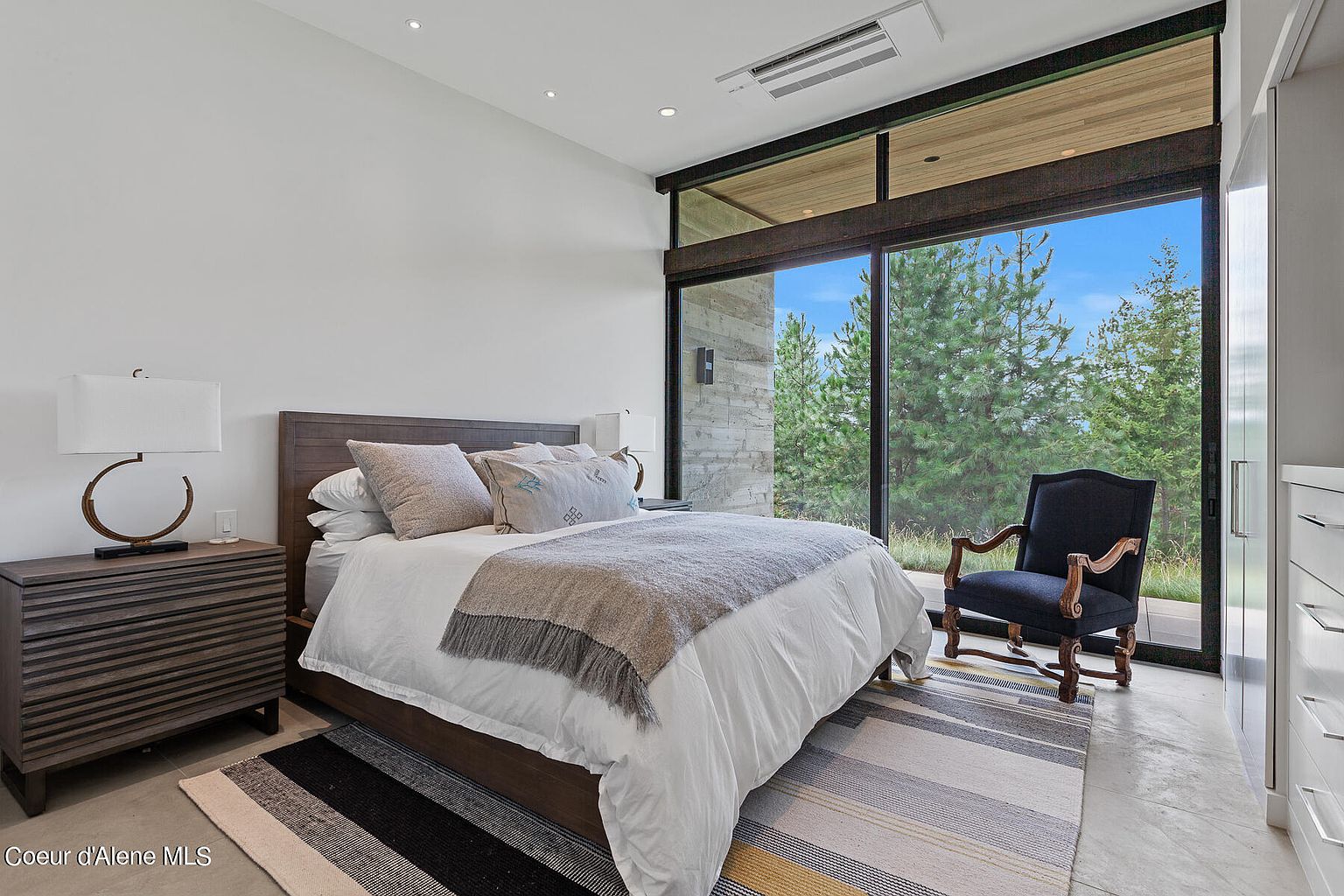 This is a primary bedroom featuring a large window with views of the surrounding trees. The room is decorated in neutral tones with a wooden bed frame, a striped rug, and a comfortable armchair. The overall impression is one of tranquility and connection with nature.