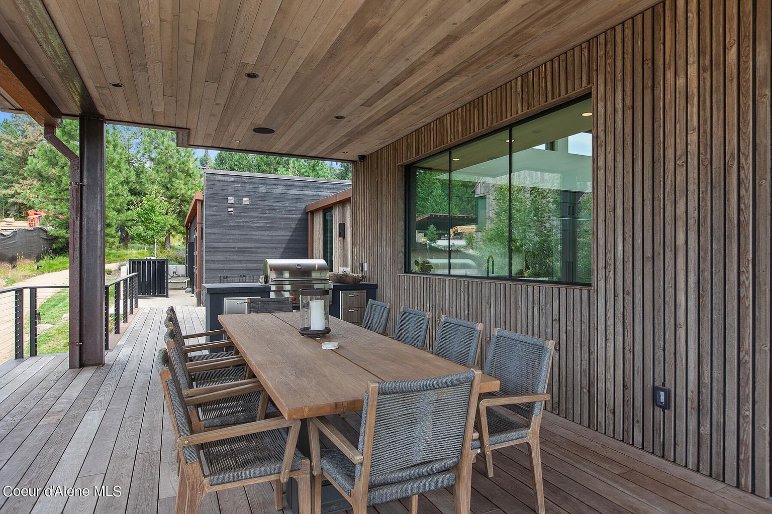 This image showcases a beautifully designed outdoor dining area on a wooden deck. The space features a long wooden table surrounded by eight woven chairs, perfect for entertaining. The deck is partially covered by a wooden ceiling, and a large window offers views of the surrounding landscape, creating a seamless blend of indoor and outdoor living.