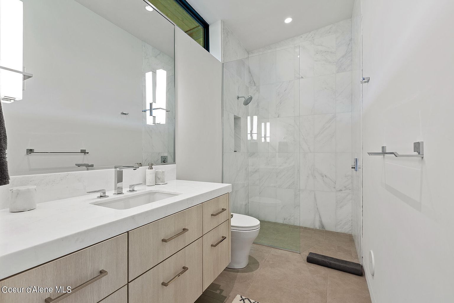 This is a primary bathroom featuring a modern vanity with light wood cabinets and a white countertop. A large mirror reflects the space, and a glass-enclosed shower with marble tile is visible. The overall design is clean and contemporary, creating a spa-like atmosphere.