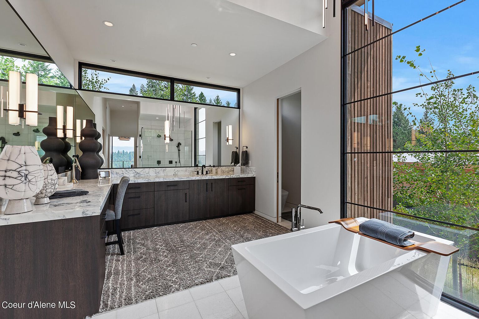 This is a luxurious primary bathroom featuring a modern freestanding bathtub positioned in front of a large window with a view of the outdoors. The bathroom includes a spacious vanity with dark wood cabinetry, a marble countertop, and a large mirror. The overall design is contemporary and elegant, emphasizing natural light and a spa-like atmosphere.