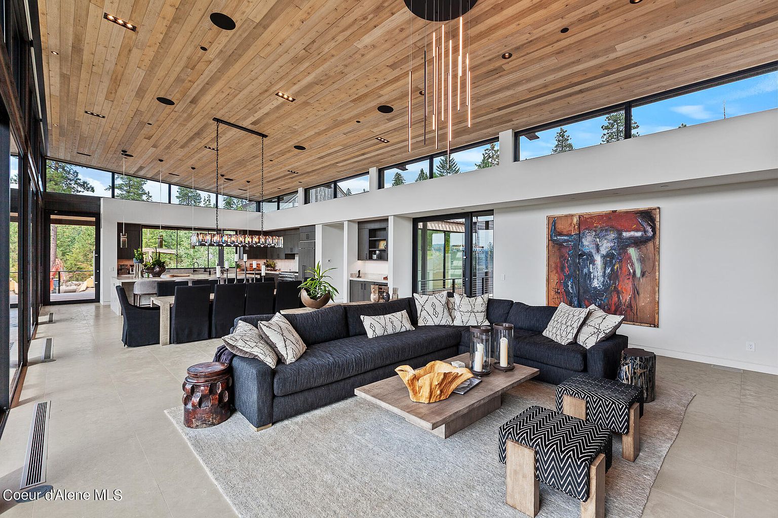 This is a spacious and modern living room featuring a large sectional sofa, a wooden coffee table, and a striking piece of art on the wall. The room has a high, wood-paneled ceiling with recessed lighting and large windows that provide ample natural light. The open floor plan seamlessly connects the living area to the dining room and kitchen, creating a cohesive and inviting space.