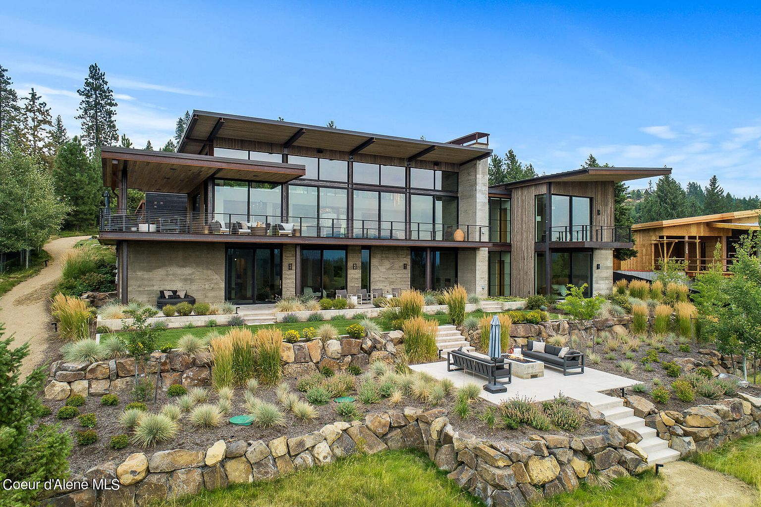 This is a striking front view of a modern, multi-level home. The architecture features a combination of concrete, wood, and expansive glass windows, creating a sophisticated and open design. The property is landscaped with terraced rock gardens and ornamental grasses, adding to the home's curb appeal and natural setting.