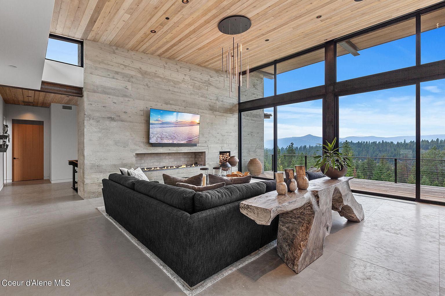 This is a luxurious living room featuring a large sectional sofa, a modern fireplace, and a wall-mounted television. The room has floor-to-ceiling windows offering a stunning view of the surrounding landscape. The decor includes natural wood elements and stone accents, creating a sophisticated and inviting atmosphere.