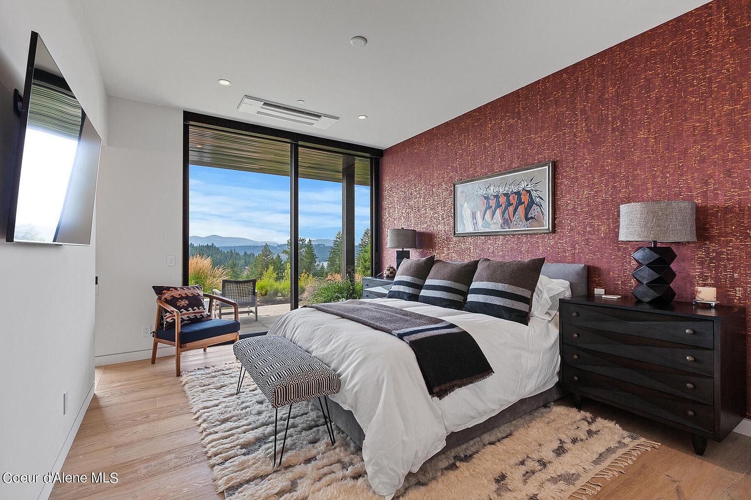 This is a primary bedroom featuring a king-size bed with a white comforter and decorative pillows. The room has a large window with a view of a lake and mountains, and a textured accent wall with artwork. The furniture includes a dark wood dresser and a bench at the foot of the bed, complemented by a plush area rug.