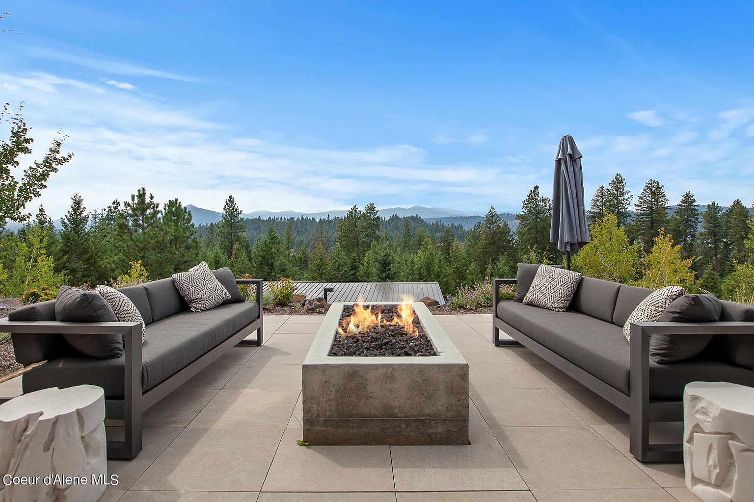 This image showcases a luxurious outdoor patio area, featuring two modern gray sofas flanking a rectangular concrete fire pit with visible flames. The patio is paved with large, light-colored tiles, and the background offers a stunning view of a forested landscape under a clear blue sky. The scene evokes a sense of relaxation and upscale outdoor living.