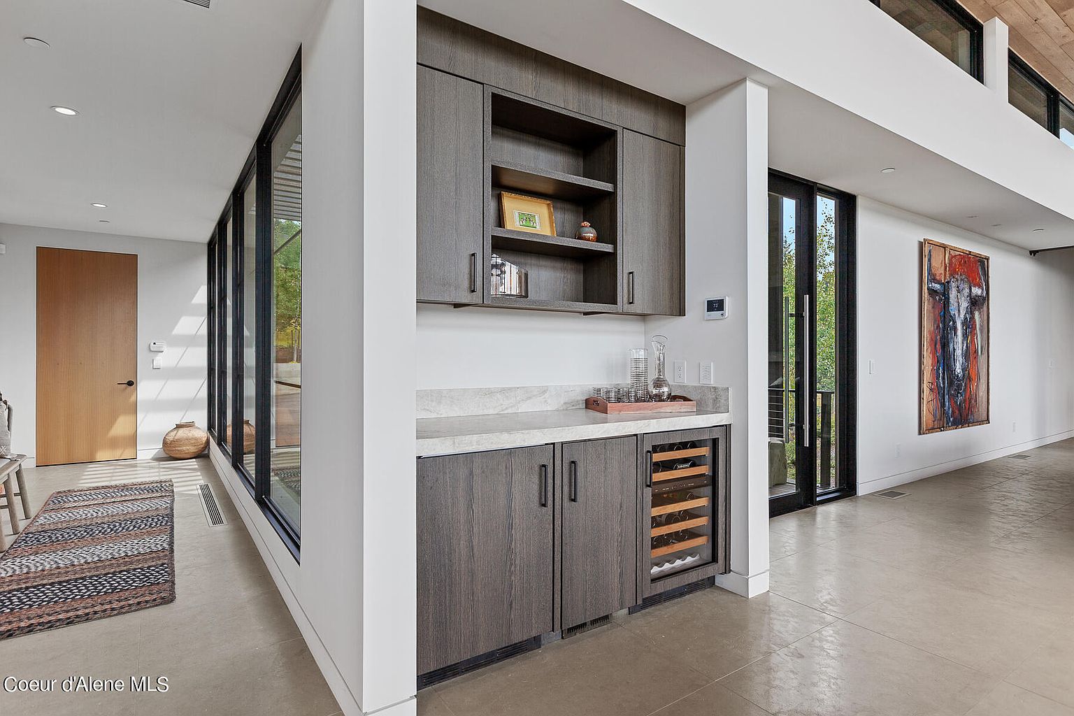 This interior shot showcases a modern hallway with a built-in bar area featuring dark wood cabinetry, a wine cooler, and a light countertop. Large windows provide natural light, and an abstract painting adds a touch of color to the white walls. The flooring appears to be polished concrete, contributing to the contemporary aesthetic.