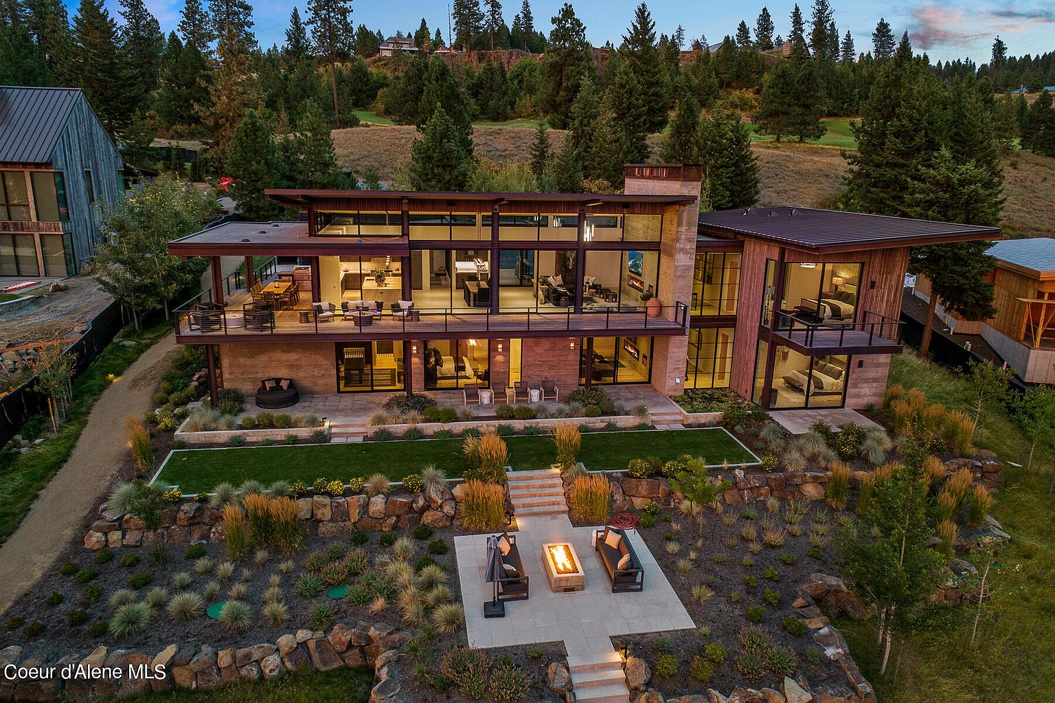 This is an aerial rear view of a modern, multi-story home with expansive windows and outdoor living spaces. The property features a well-manicured lawn, tiered landscaping with rock features, and a fire pit area with seating. The architecture emphasizes clean lines and a seamless integration with the natural surroundings, creating a luxurious and inviting atmosphere.