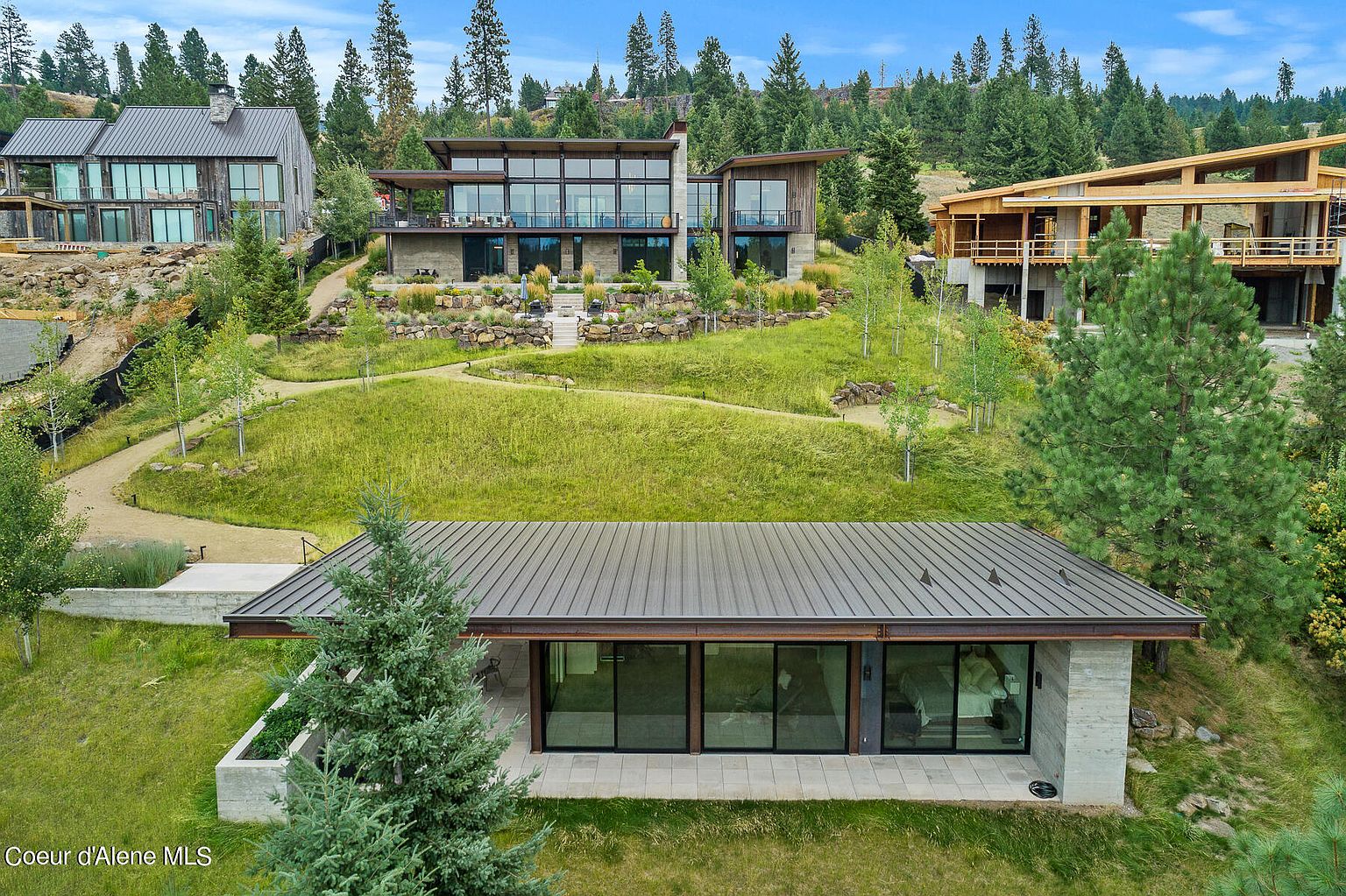 This aerial view showcases a modern luxury home nestled on a lush, green hillside. The property features multiple structures with contemporary architectural designs, including clean lines, large windows, and metal roofing. The landscape is meticulously maintained with manicured lawns, mature trees, and stone pathways, creating a serene and upscale living environment.