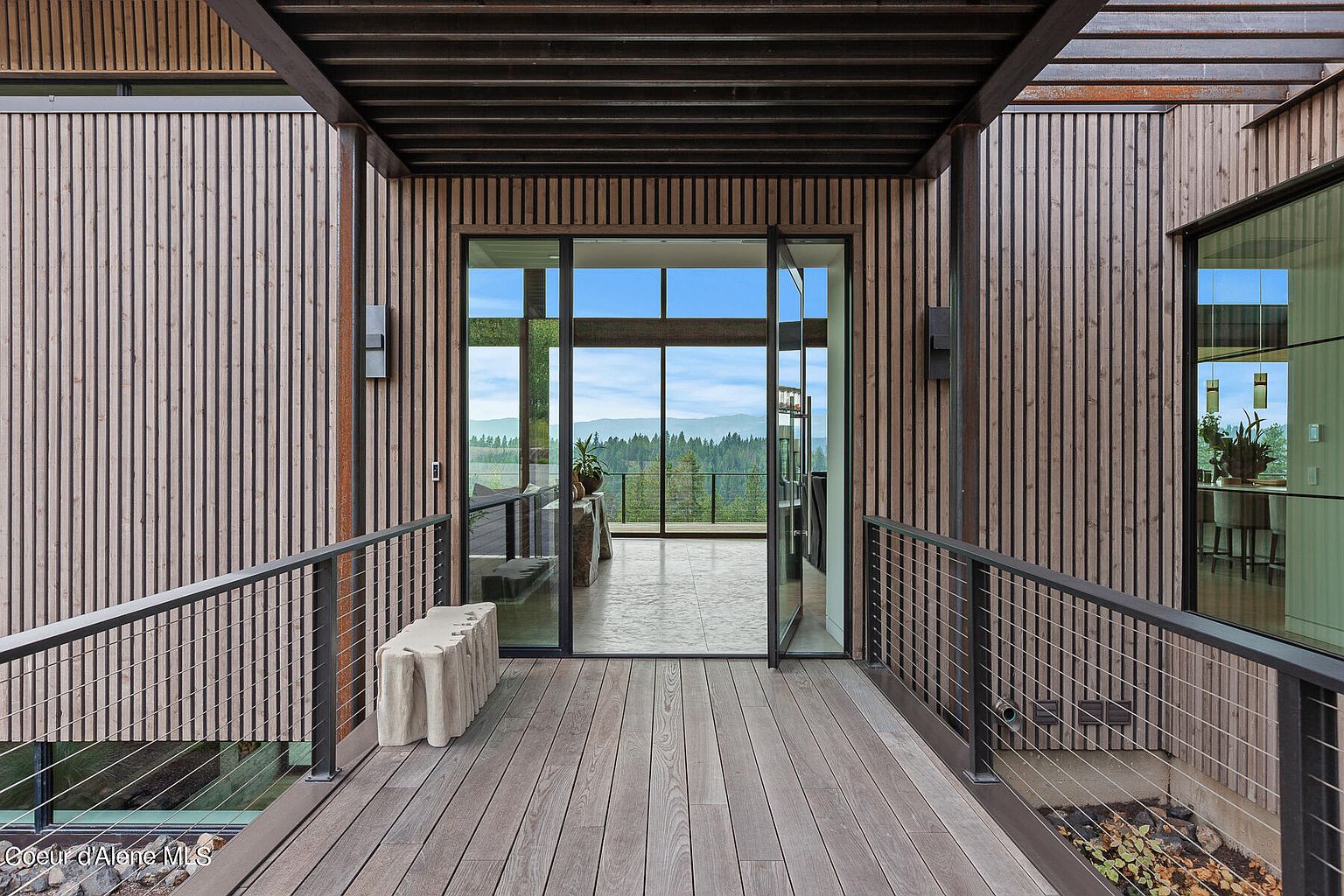 This image showcases a modern patio or deck area with a stunning view of the mountains and forest. The space features wooden decking, a metal railing with cable wires, and a unique stone bench. The design emphasizes clean lines and natural materials, creating a serene and inviting outdoor living space.