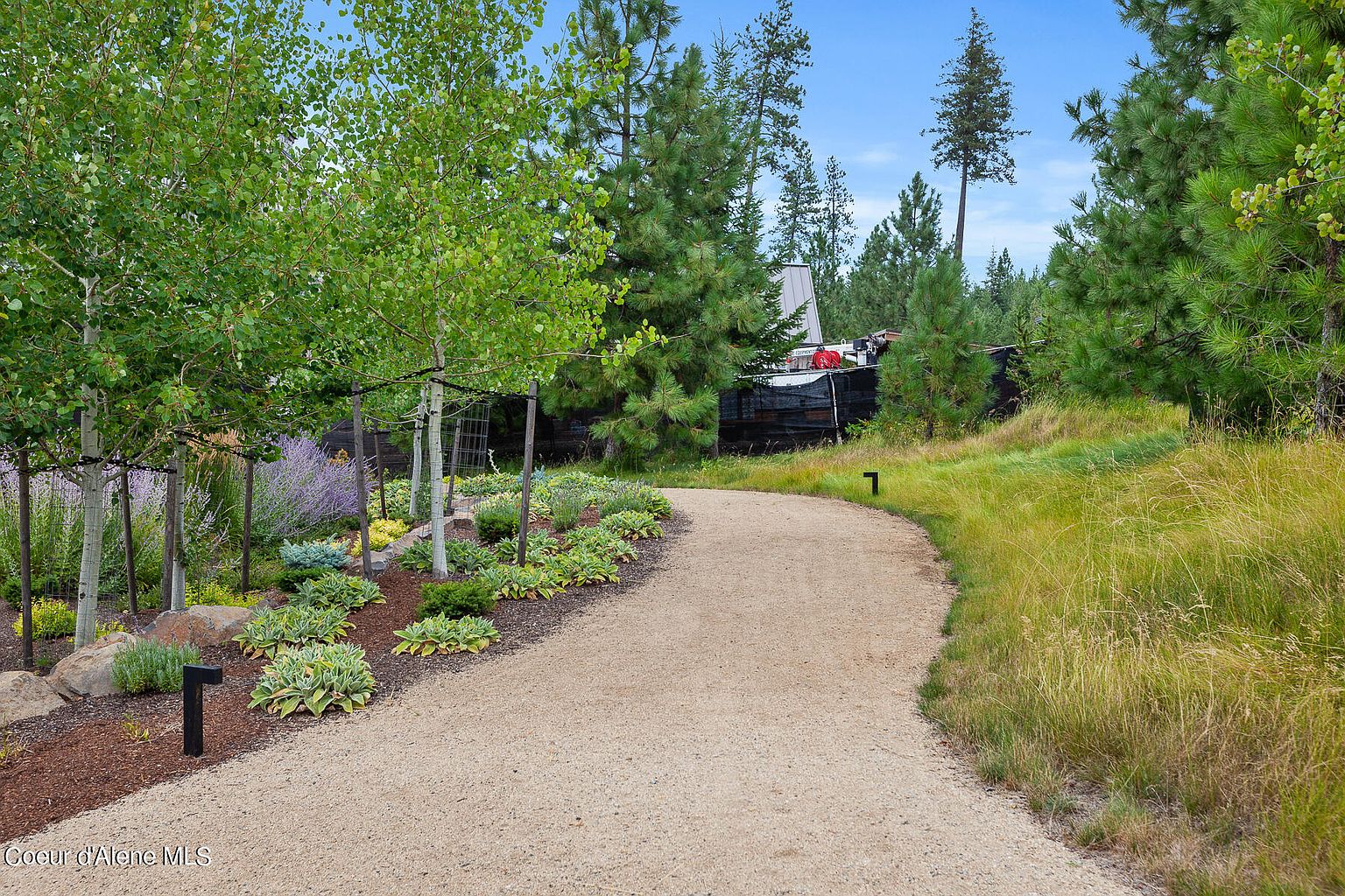 This image showcases a beautifully landscaped yard with a gravel pathway winding through lush greenery. The garden features a variety of plants, including trees, shrubs, and flowering perennials, creating a serene and inviting atmosphere. The pathway leads towards a partially obscured building, suggesting a well-maintained and private property.