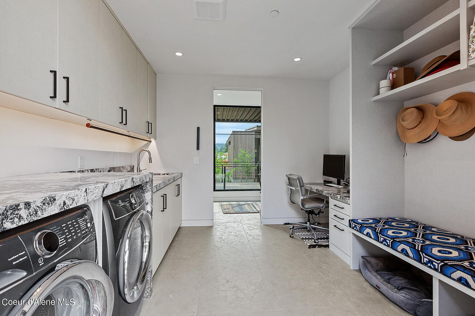 This is a well-organized laundry room featuring modern cabinetry, granite countertops, and front-loading washer and dryer. The room also includes a built-in bench with storage, a small desk area, and access to an outdoor balcony. The overall impression is clean, functional, and stylish.