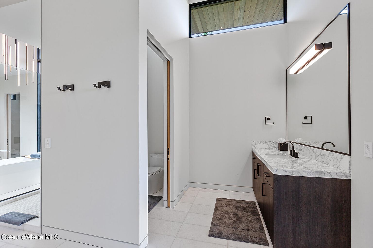 This is a bright and modern primary bathroom. It features a vanity with a marble countertop and dark wood cabinetry, a large mirror with integrated lighting, and a separate toilet room. The walls are painted white, and the floor is tiled, creating a clean and spacious feel.