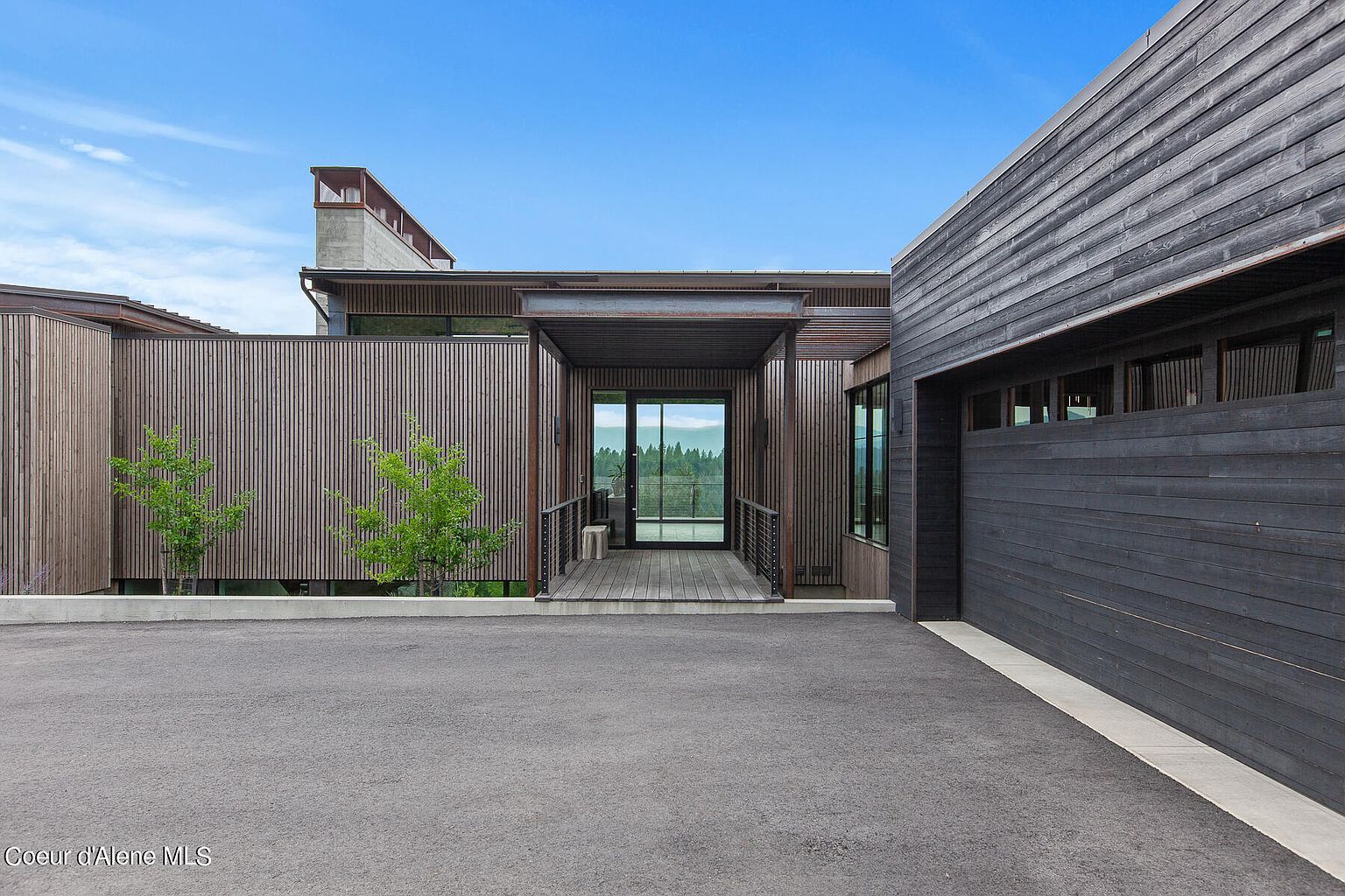 This image showcases the entryway of a modern home, featuring a covered walkway leading to a glass-door entrance. The exterior is clad in wood siding, with a combination of vertical and horizontal planks, and a dark-colored garage door is visible on the right. The asphalt driveway provides ample parking space, and the overall design exudes a contemporary and sophisticated aesthetic.