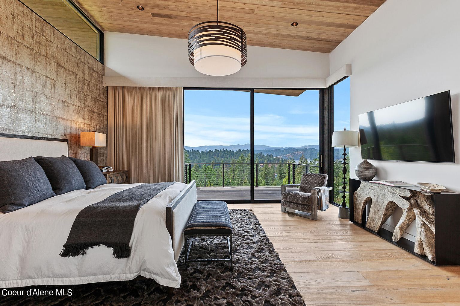 This is a luxurious primary bedroom featuring a king-size bed with plush bedding, a textured accent wall, and a stunning view through large sliding glass doors leading to a balcony. The room is decorated in a modern style with a unique light fixture, a comfortable armchair, and a custom-designed console table, creating a serene and upscale atmosphere.