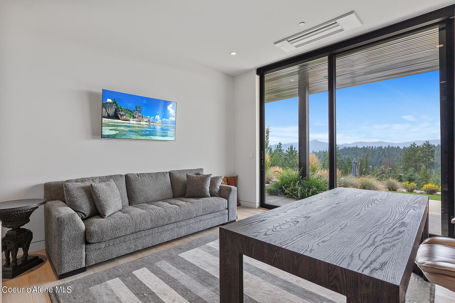 This interior shot showcases a modern living room with a gray sofa, a large flat-screen TV displaying a beach scene, and a dark wood table. Large sliding glass doors offer a stunning view of the landscape outside, bringing natural light into the room. The room is decorated in neutral tones, creating a serene and inviting atmosphere.