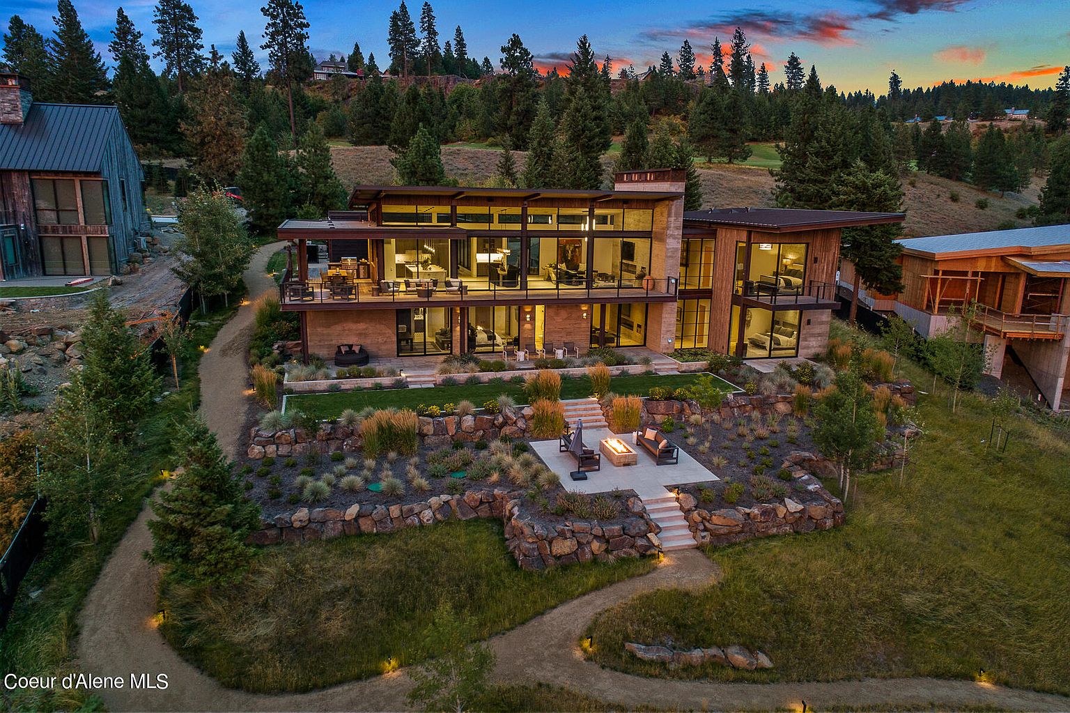 This is an aerial rear view of a modern, multi-story home with large windows and a tiered backyard. The property features a stone-lined garden, a fire pit area with seating, and a winding pathway leading to the house. The surrounding landscape includes mature trees and a glimpse of a golf course, creating a serene and luxurious atmosphere.