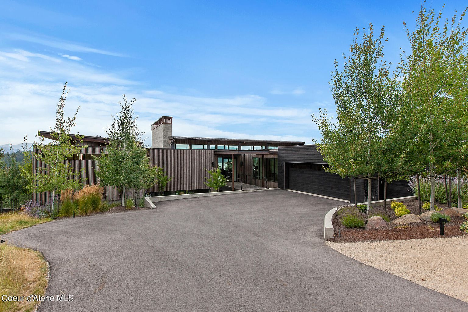 This is a front exterior view of a modern home with a long, horizontal design. The house features a mix of wood siding and a flat roof, complemented by a concrete driveway. Landscaping includes trees and gravel beds, adding to the property's curb appeal.