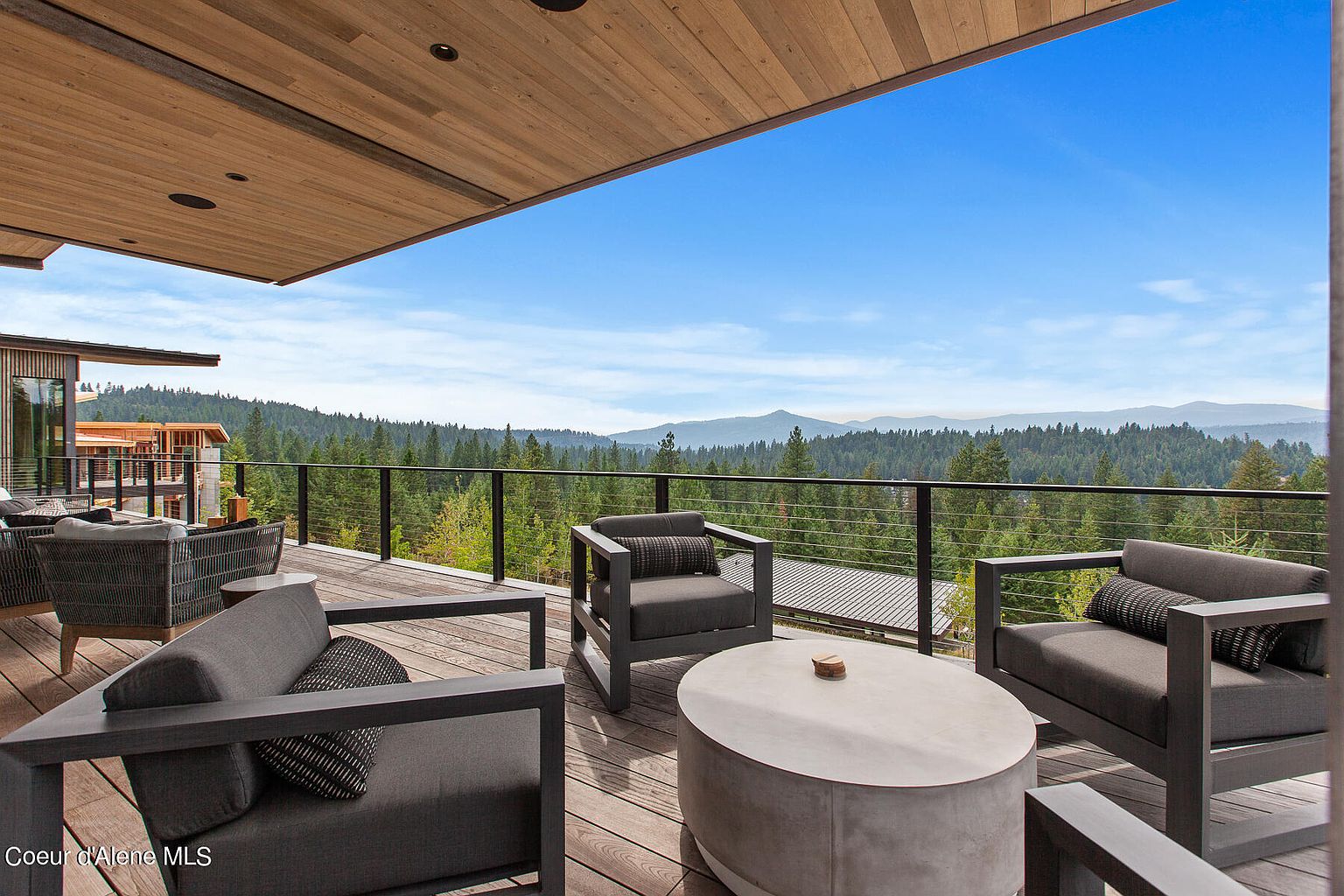 This image showcases a luxurious outdoor deck area with modern furnishings, including gray armchairs and a round concrete coffee table. The deck is constructed with wooden planks and features a sleek railing system, offering an unobstructed view of the surrounding forest and distant mountains under a clear blue sky. The covered ceiling adds a touch of sophistication and protection from the elements.
