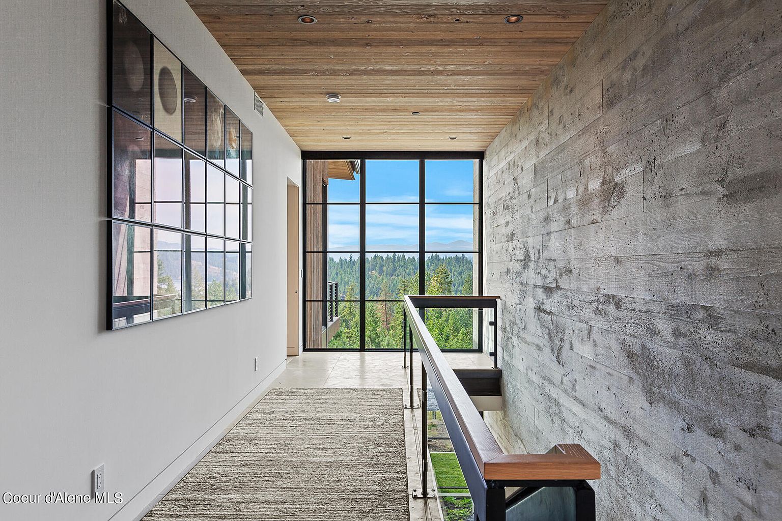 This interior shot showcases a modern hallway with a striking combination of textures and natural light. A large window at the end of the hallway provides a view of the surrounding landscape, while a concrete wall adds an industrial touch. The hallway features a wooden ceiling, a neutral-toned rug, and a staircase with a glass railing, creating a sophisticated and inviting space.