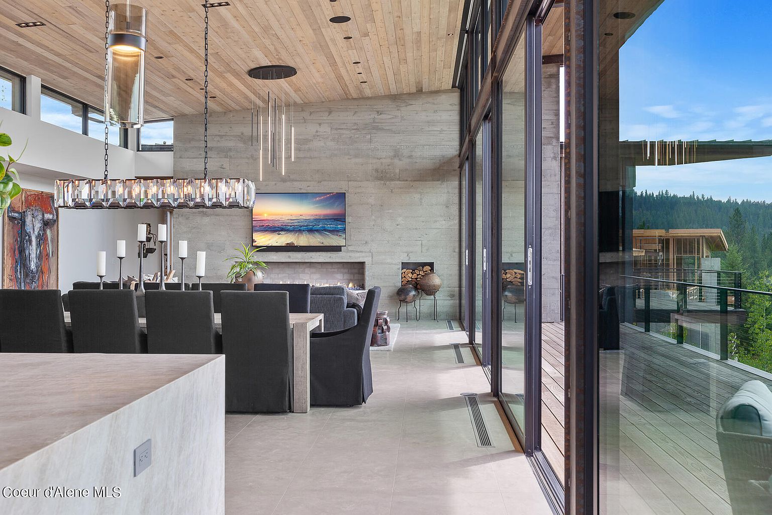 This is a modern dining room featuring a long wooden table with upholstered chairs under a crystal chandelier. The room has a concrete accent wall with a fireplace and a large television above it. Floor-to-ceiling windows offer a view of the outdoor deck and surrounding landscape, creating a seamless indoor-outdoor living experience.