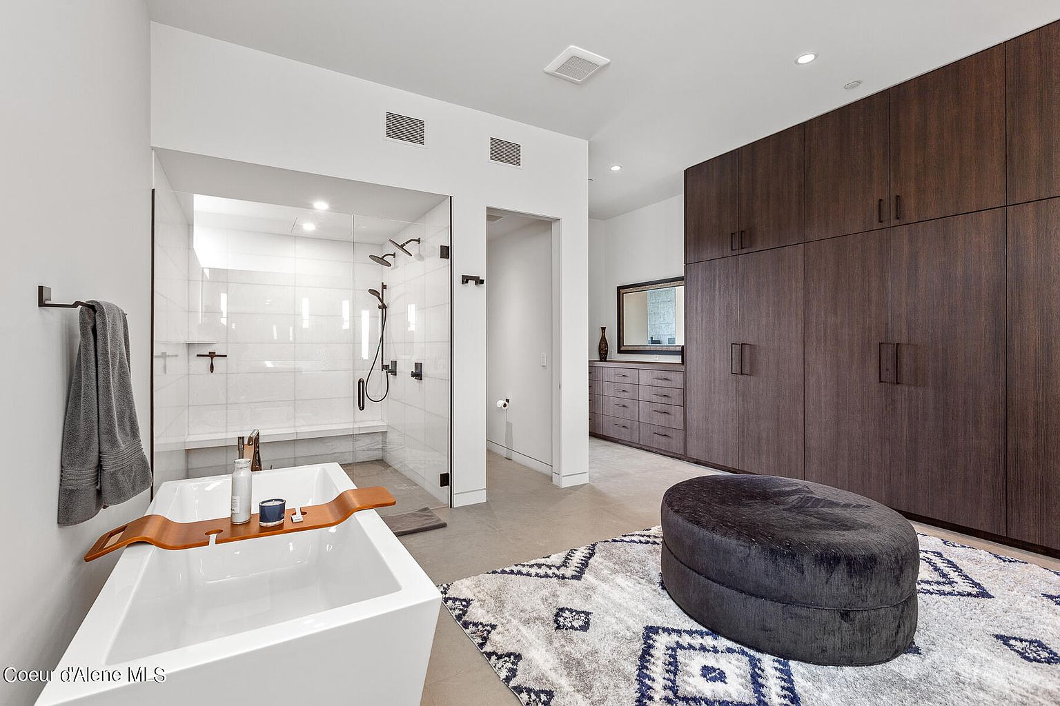 This is a luxurious primary bathroom featuring a modern freestanding bathtub with a wooden tray, a glass-enclosed shower with dual showerheads, and a large dark wood wardrobe. The bathroom also includes a gray patterned rug and a dark blue ottoman, adding a touch of elegance and comfort. The overall impression is clean, spacious, and upscale.