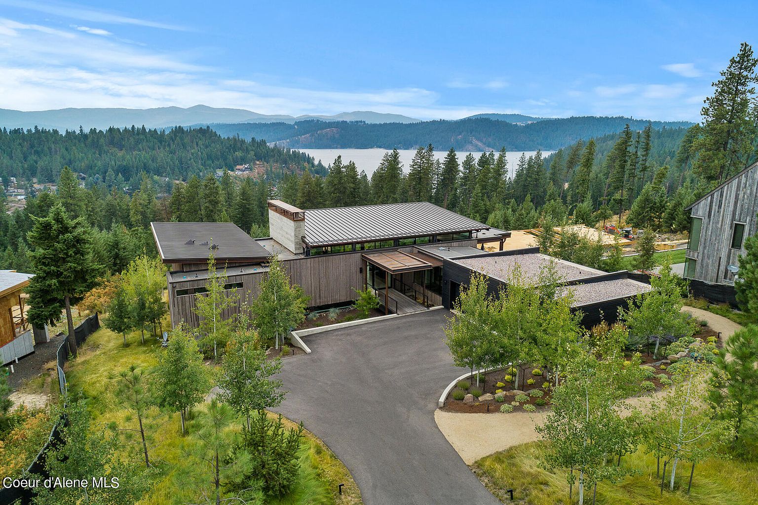 This aerial view showcases a modern, architecturally striking home nestled amidst a lush forest setting with a lake in the background. The property features a long driveway leading to the house, a combination of flat and sloped roofs, and a blend of wood and dark-colored exterior finishes. The landscaping is natural and integrates seamlessly with the surrounding environment, offering both privacy and stunning views.