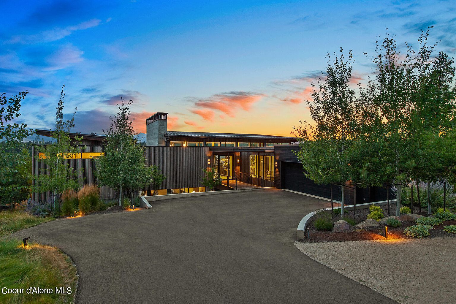 This image showcases the front exterior of a modern home at dusk. The house features a combination of wood and concrete elements, with large windows and a flat roofline. A paved driveway leads up to the entrance, and the landscaping includes trees, shrubs, and decorative lighting, creating an inviting and sophisticated curb appeal.