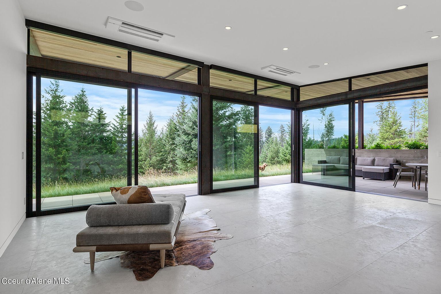 This is a bright and spacious living room with floor-to-ceiling windows that offer stunning views of the surrounding forest. The room features a modern sofa, a cowhide rug, and a minimalist design. The open floor plan seamlessly connects the interior with an outdoor patio, creating an inviting and luxurious living space.